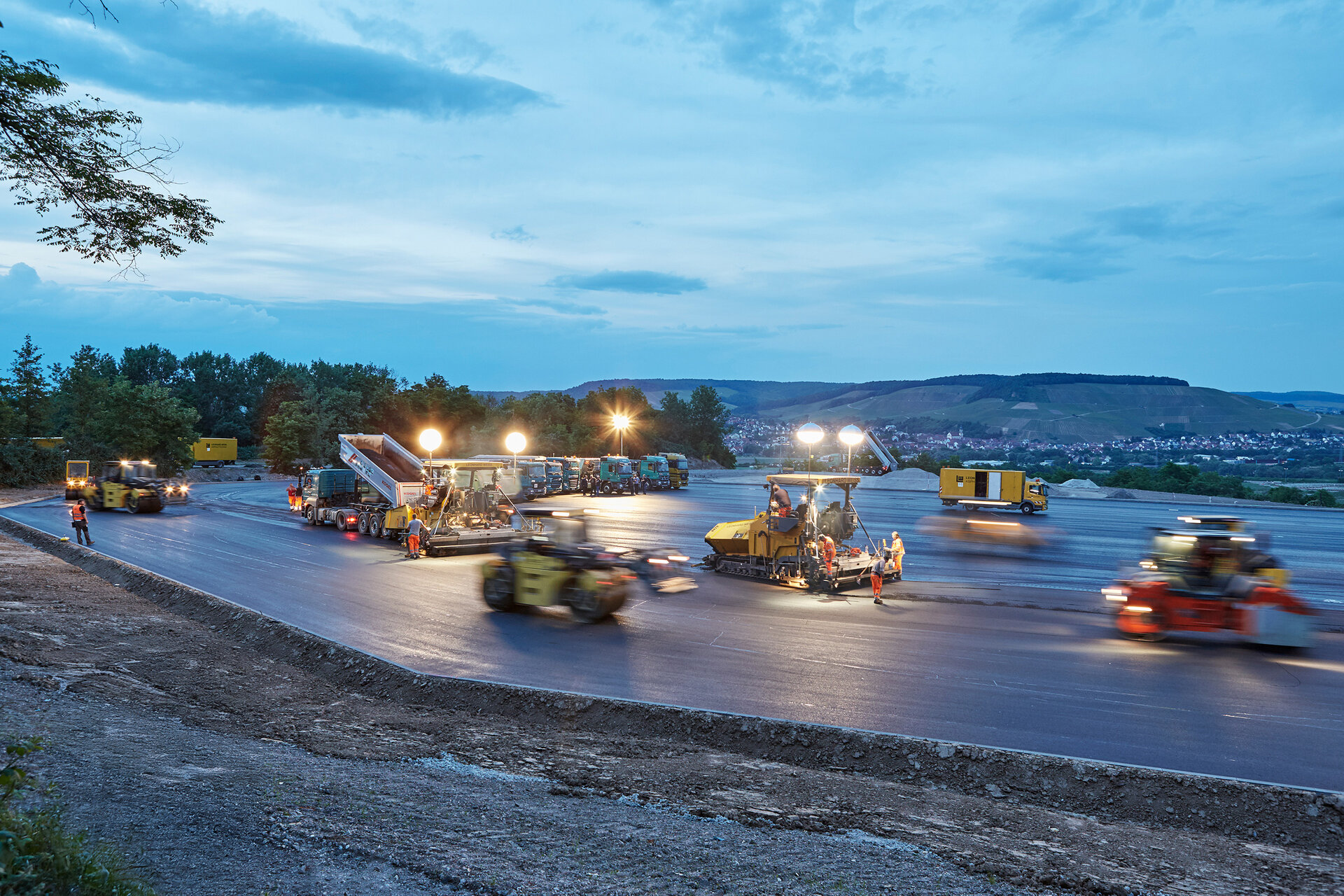 Bei Dämmerung arbeiten Bauarbeiter an einer neuen Straße, umgeben von schweren Maschinen und hellen Scheinwerfern.