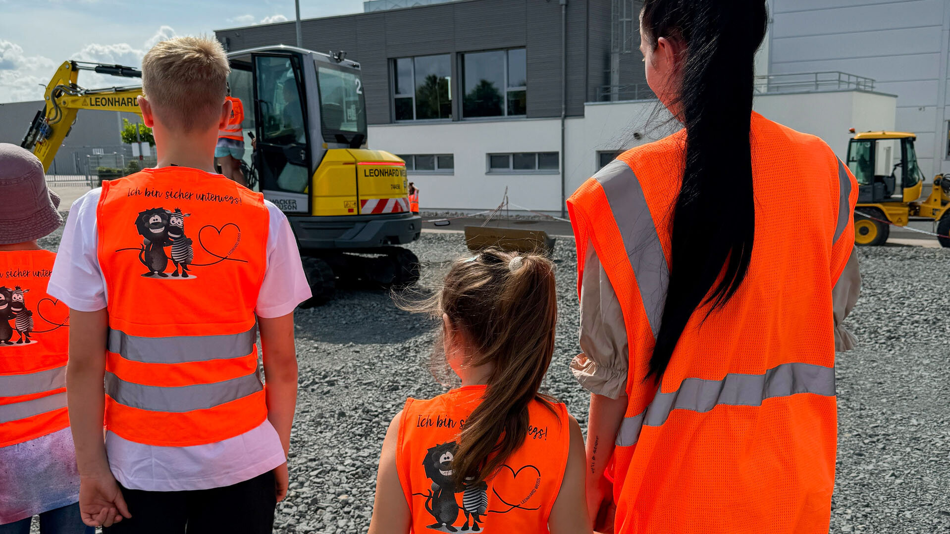 Eine Gruppe von Kindern in leuchtend orangefarbenen Sicherheitswesten steht auf einem Kiesplatz und beobachtet Bagger.