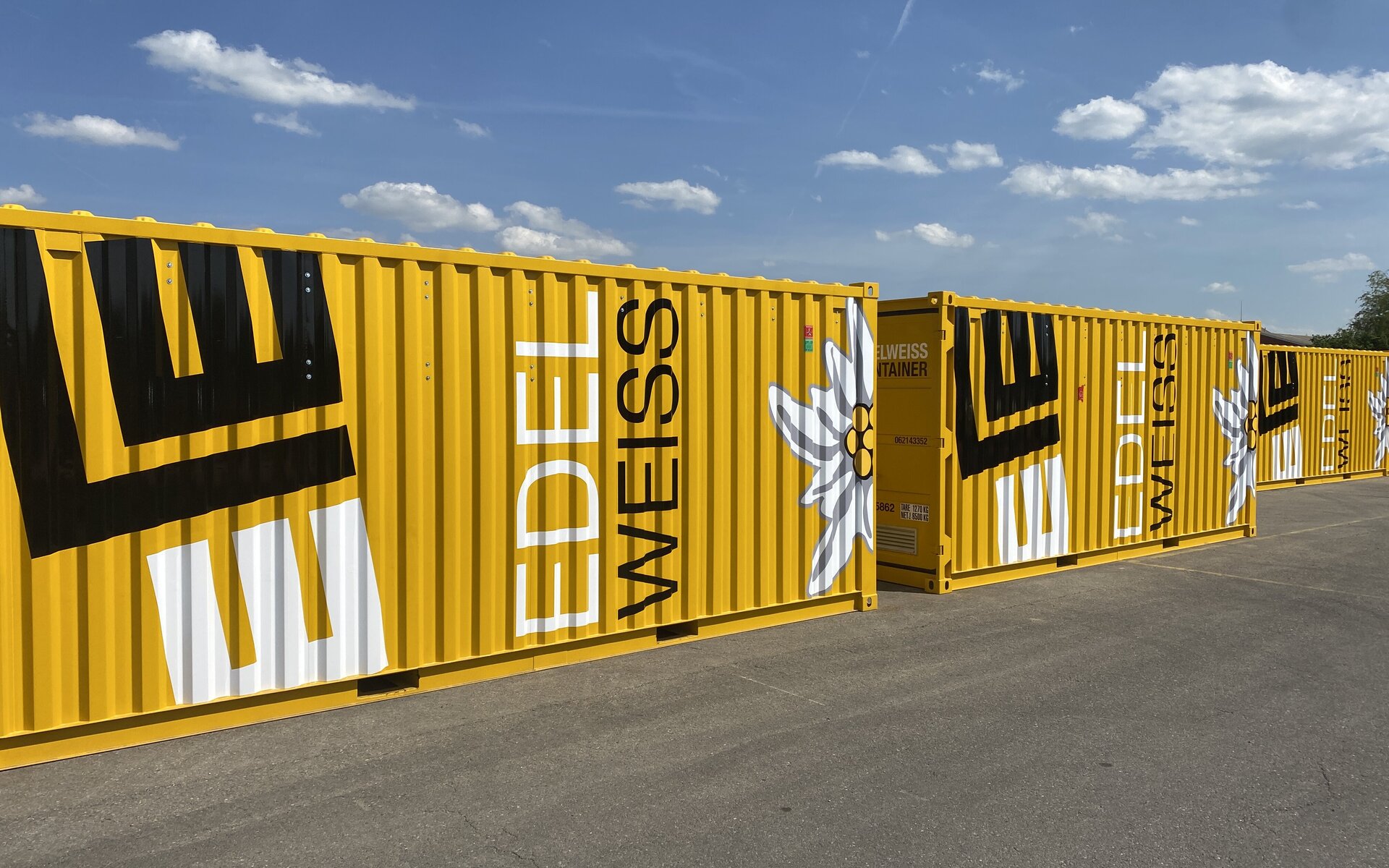 Die gelben Container sind mit dem Schriftzug 'EDELWEISS' und einer stilisierten Edelweißblume dekoriert, unter einem blauen Himmel.