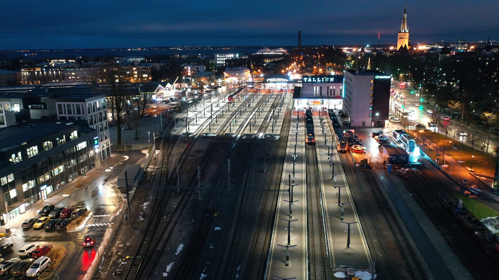 A night-time view of Tallinn railway station, illuminated by street lamps and surrounded by modern buildings.