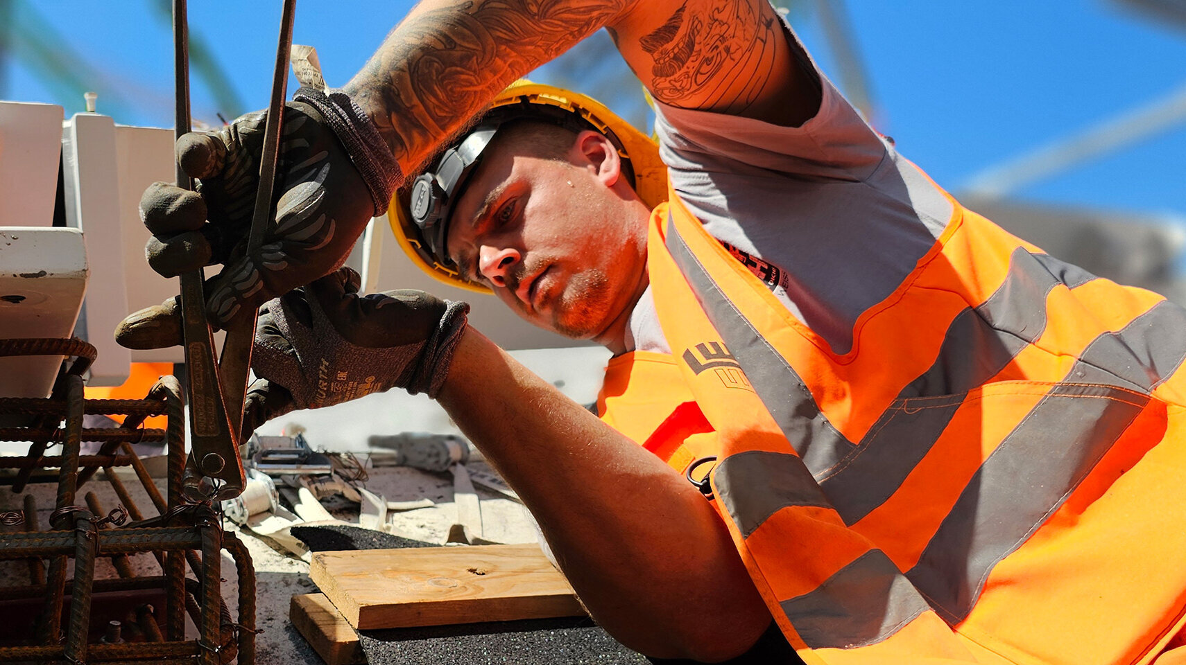 Ein Bauarbeiter in orangefarbener Sicherheitskleidung bindet im Freien mit einer Zange einen Bewehrungskorb.