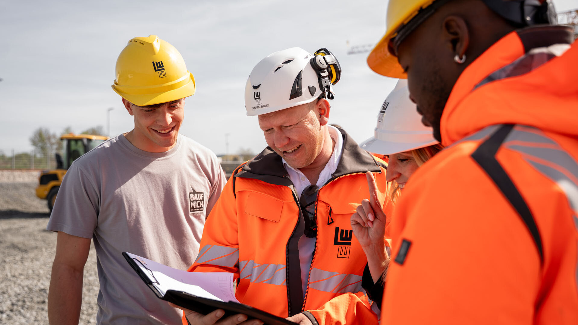 Ein Team von Bauprofis, gekleidet in leuchtend orangefarbene Jacken, analysiert gemeinsam ein Projekt auf einem sonnigen Baustellengelände.
