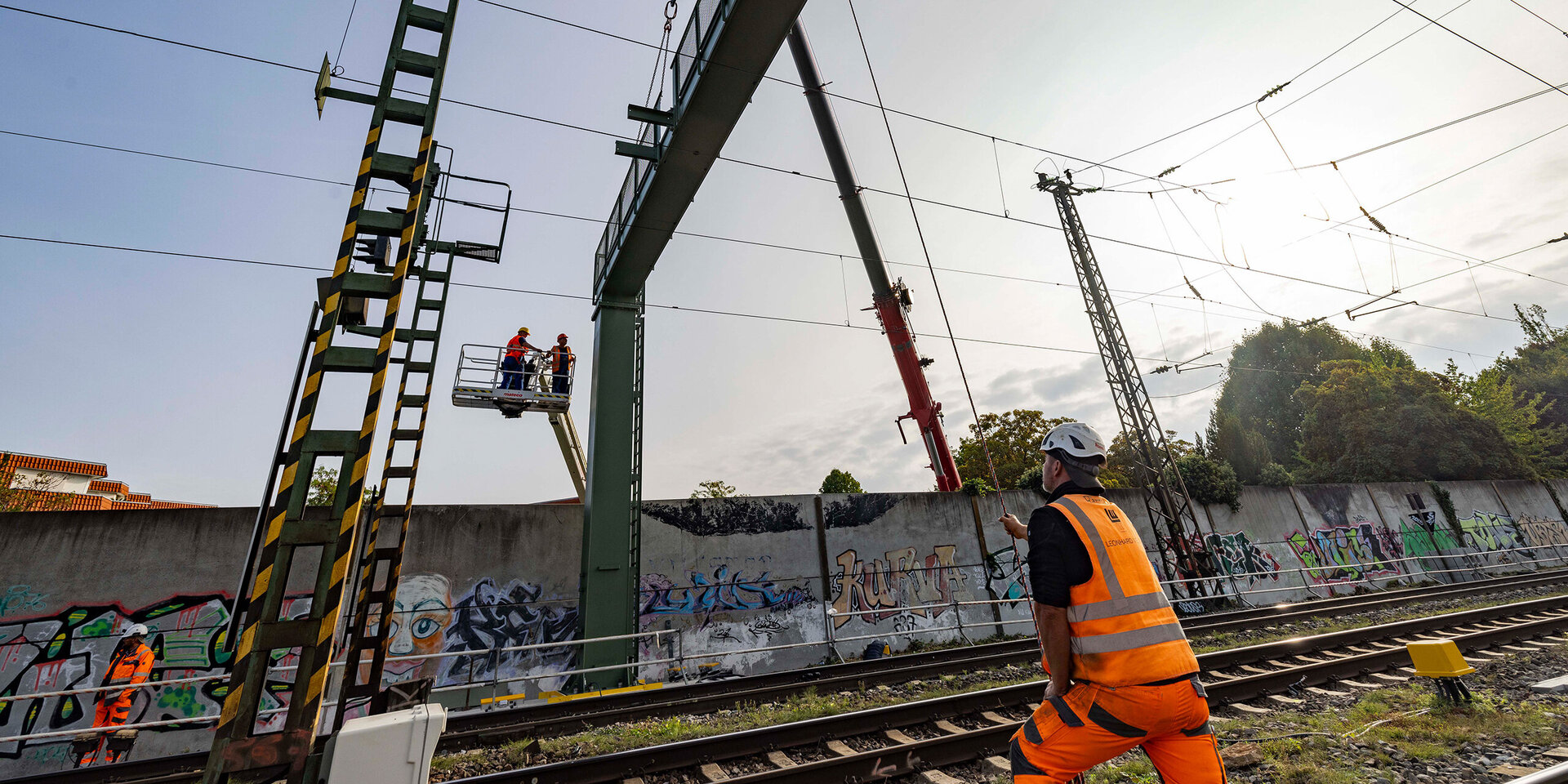 Ein Mann in Sicherheitskleidung schaut zu, während eine Hebebühne mit Arbeitern an einer hohen Struktur über den Gleisen schwebt.