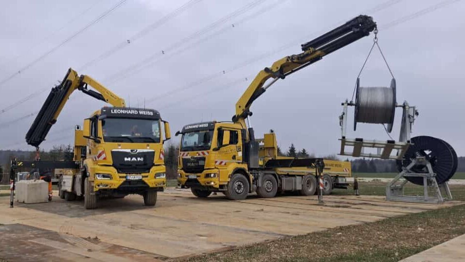 Zwei gelbe Lkw mit Kränen heben eine große Kabelrolle und Betonklötze auf einer Baustelle unter einem bewölkten Himmel.