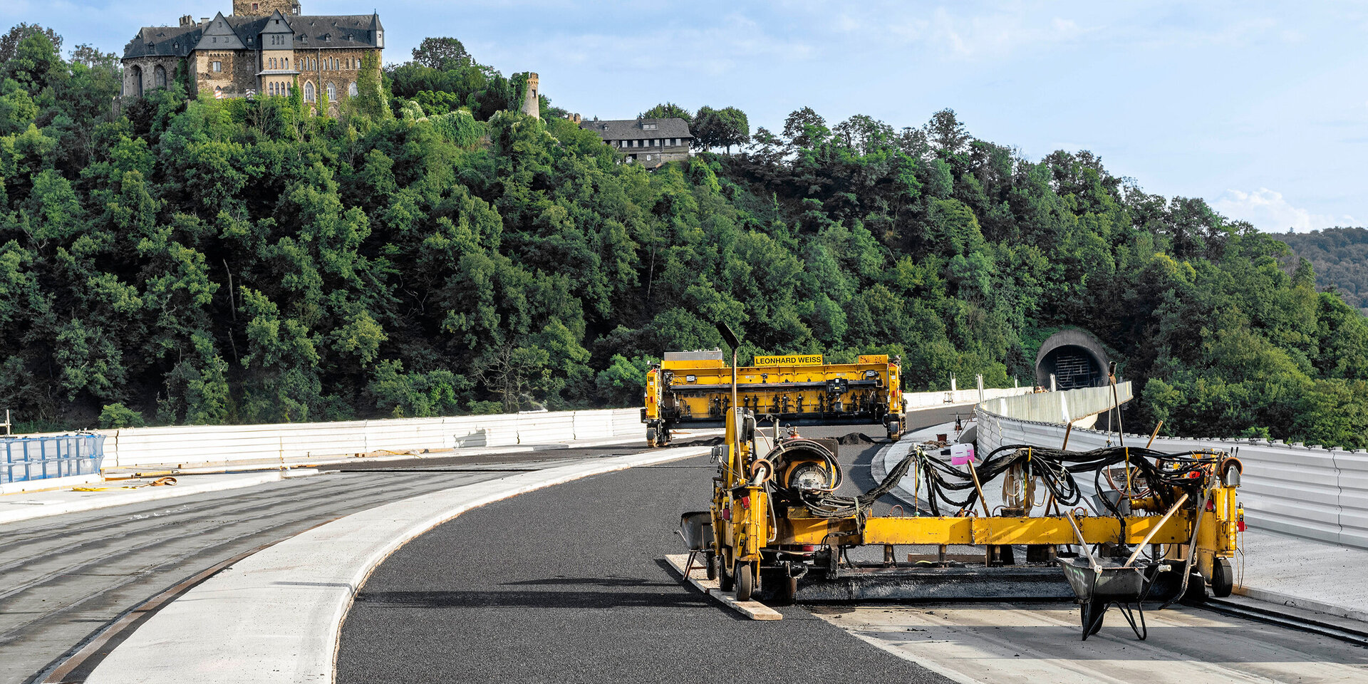 Eine Baustelle zeigt eine Gussasphalteinbaubohle, die Asphalt auf einer kurvenreichen Straße verlegen, mit einer historischen Burg im Hintergrund.
