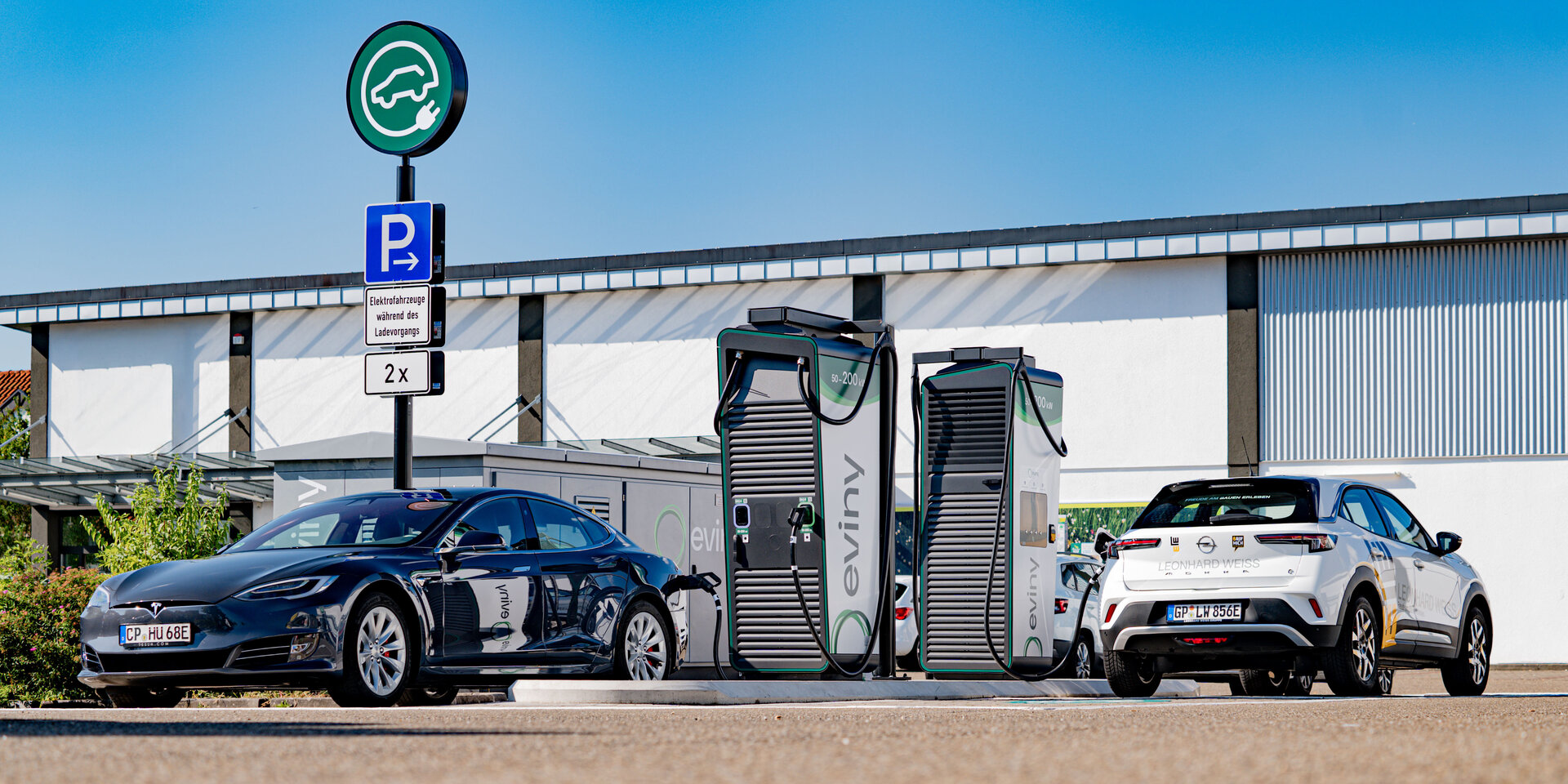 Zwei Elektroautos stehen an modernen Ladestationen, umgeben von einem klaren blauen Himmel und einer hellen Umgebung.