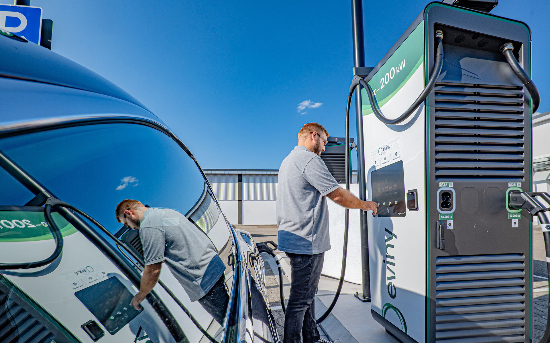 Der Fahrer eines Elektroautos interagiert mit einer modernen Ladestation, um sein Fahrzeug aufzuladen, bei klarem Himmel.