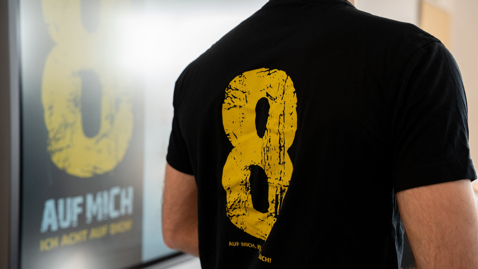 Ein Mann trägt ein schwarzes T-Shirt mit einer großen gelben Zahl 8 auf dem Rücken, während er vor einem Bildschirm steht.