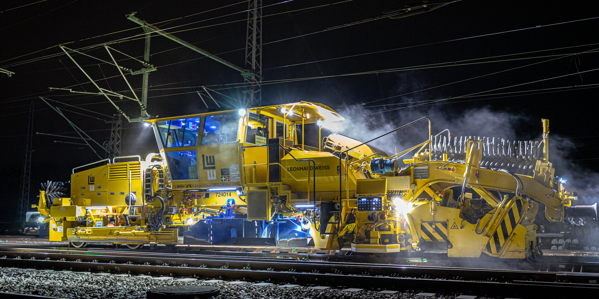 Eine gelbe Gleisbaumaschine arbeitet nachts an den Schienen, umgeben von Rauch und beleuchtet von starken Scheinwerfern.