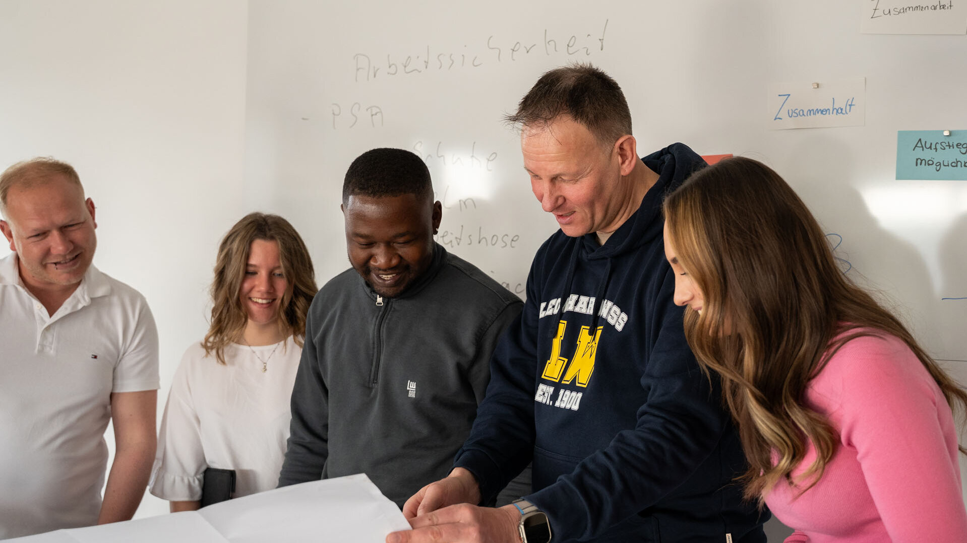 In einem hellen Raum zeigen fünf Menschen Interesse an einem Dokument, das auf dem Tisch ausgebreitet ist, und tauschen Ideen aus.