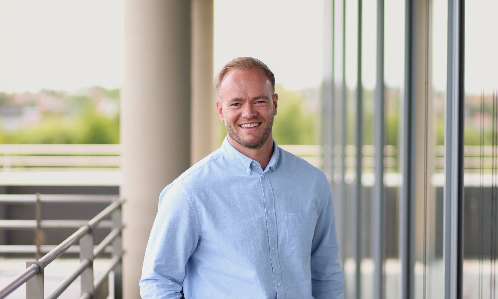 Porträtfoto eines freundlich lächelnden Mannes im hellblauen Hemd, der sich auf einer modernen Balkonterrasse mit Glasfassade und Blick ins Grüne entspannt anlehnt.