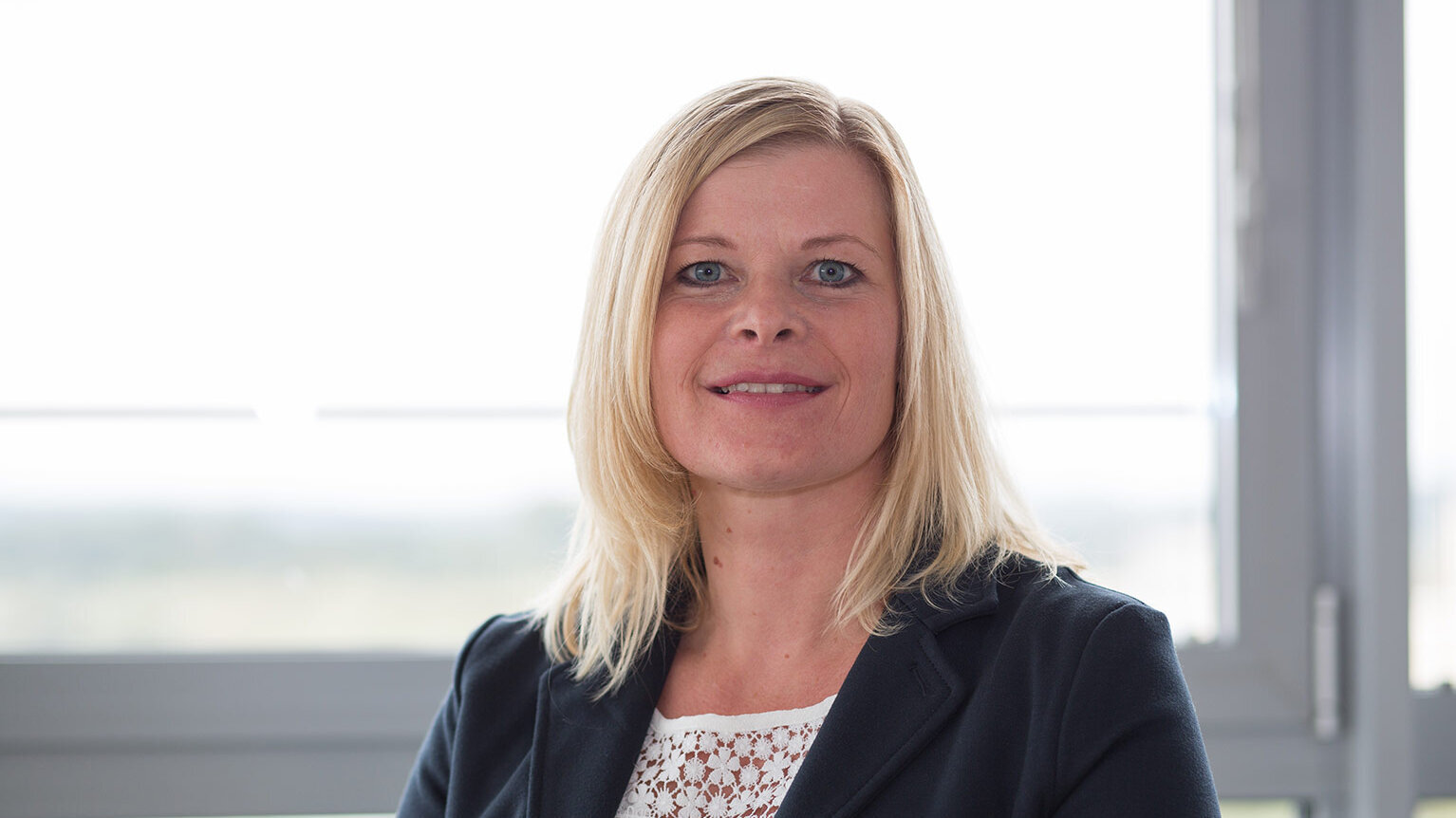 Porträt einer Frau mit schulterlangem blondem Haar, die lächelnd in die Kamera blickt. Sie trägt eine weiße Bluse mit Spitzenmuster und einen dunklen Blazer, aufgenommen in einem hellen Bürogebäude.