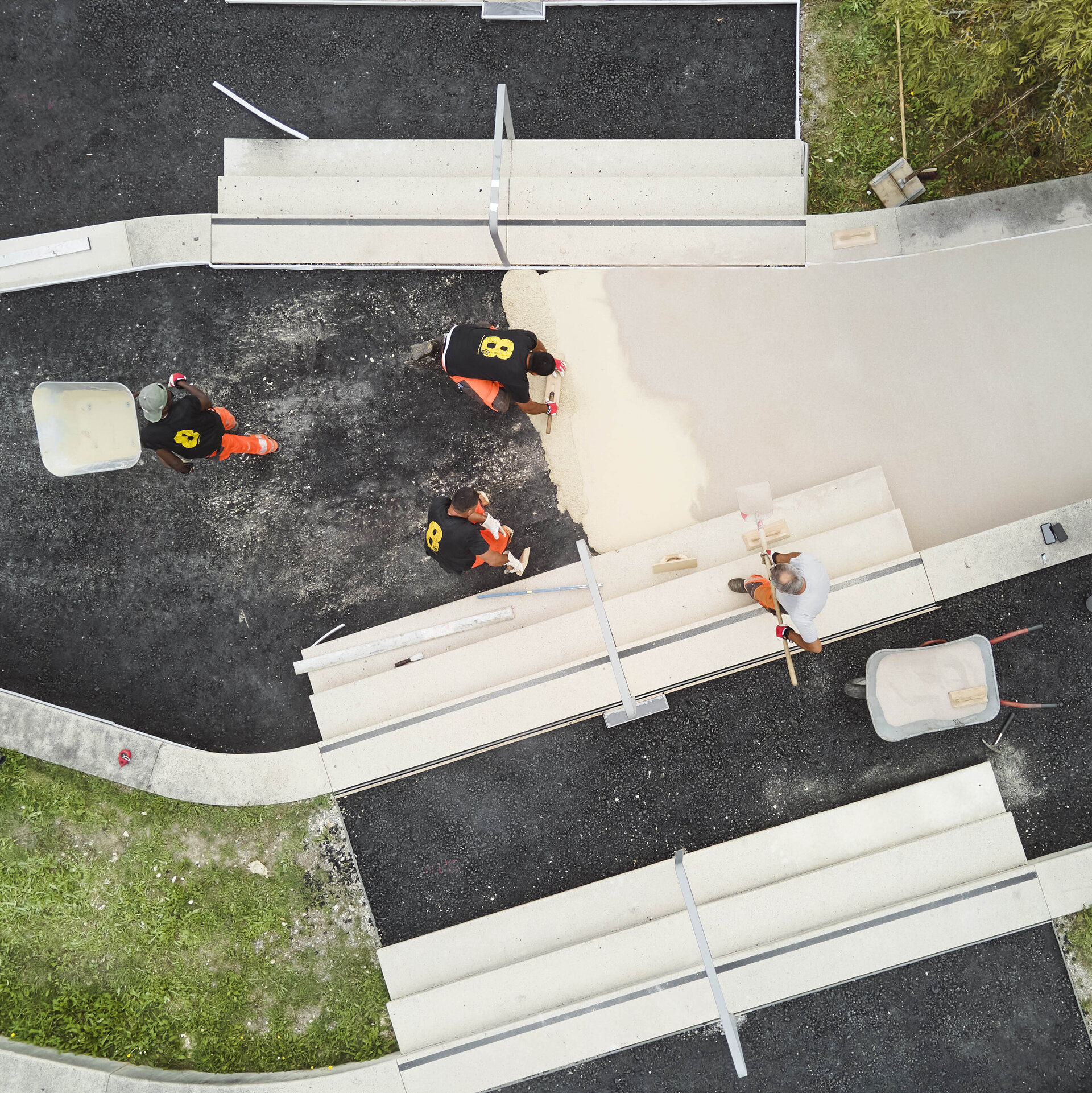 Drei Bauarbeiter in orangefarbenen Hosen arbeiten an einer Baustelle, während sie Material auf den Boden verteilen.