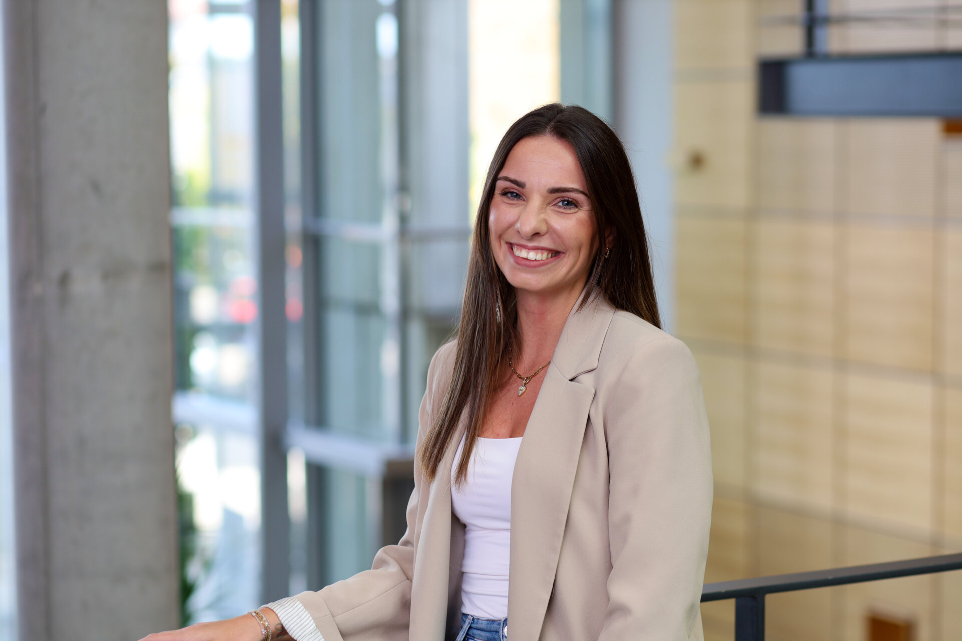 Eine junge Frau mit langen, dunklen Haaren lächelt freundlich in die Kamera, während sie in einem modernen Raum steht.