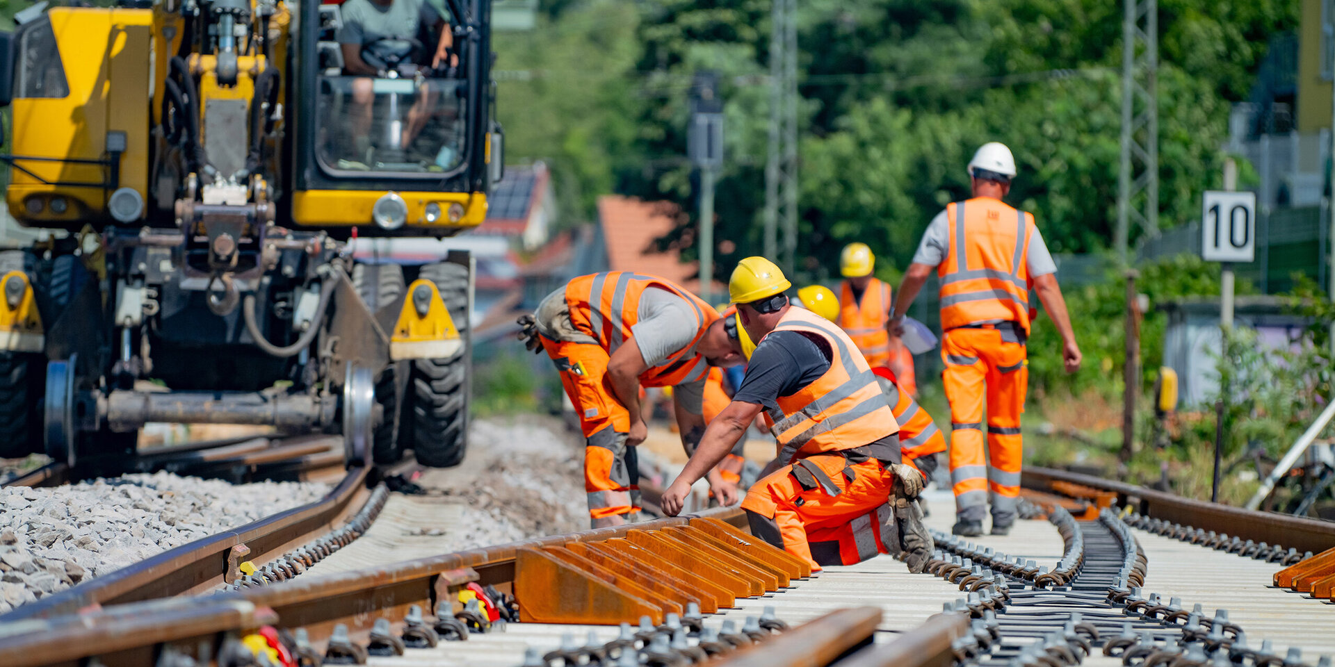 Im sonnigen Wetter arbeiten mehrere Bauarbeiter an den Gleisen, während ein gelbes Fahrzeug die Baustelle unterstützt.