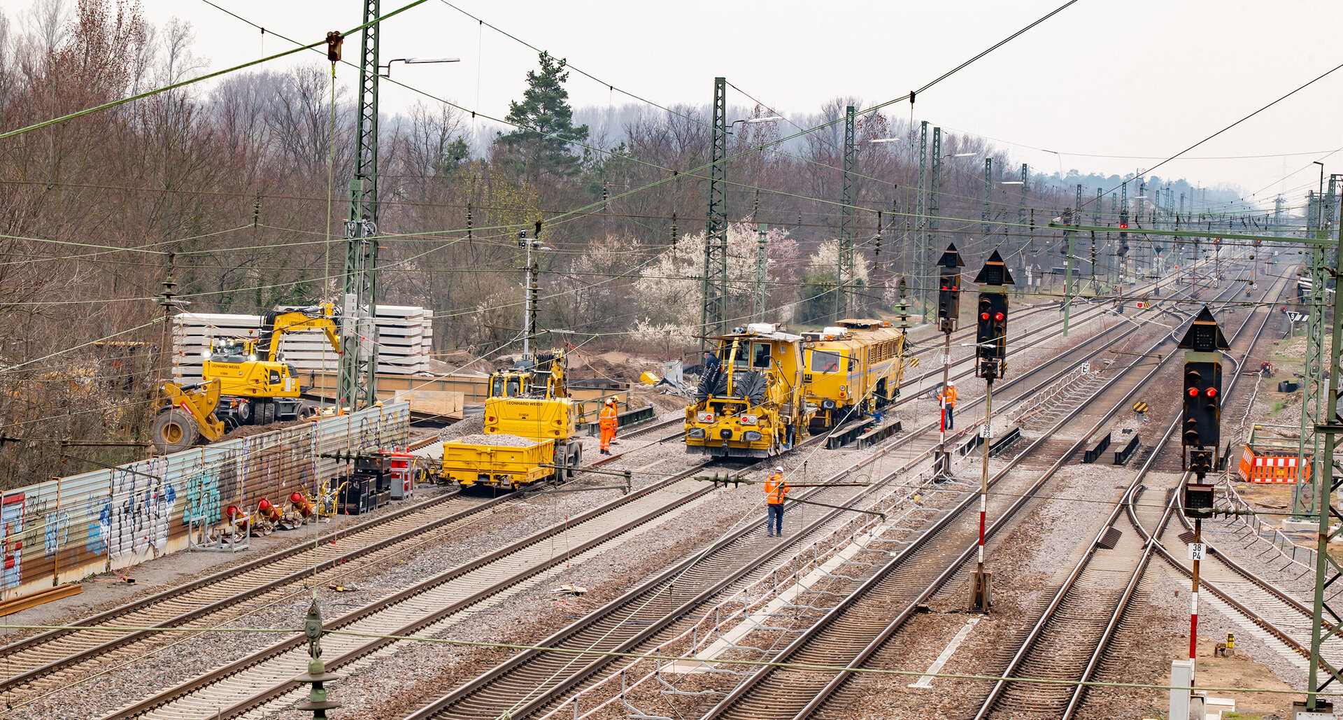 Auf einer Baustelle arbeiten mehrere gelbe Maschinen an den Gleisen, während Arbeiter in orangefarbener Kleidung aktiv sind.