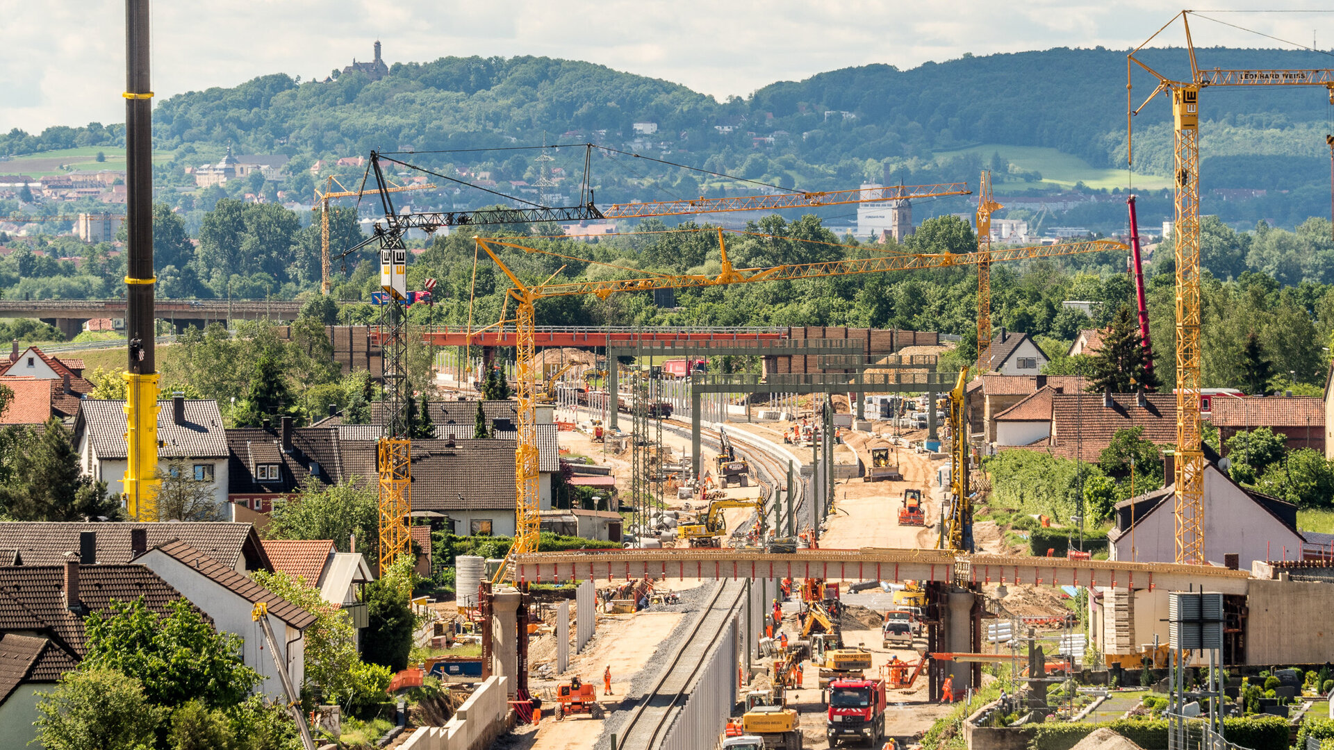 Eine Baustelle mit mehreren Kränen und Baufahrzeugen, die an einer neuen Eisenbahnlinie arbeiten, umgeben von grünen Hügeln.