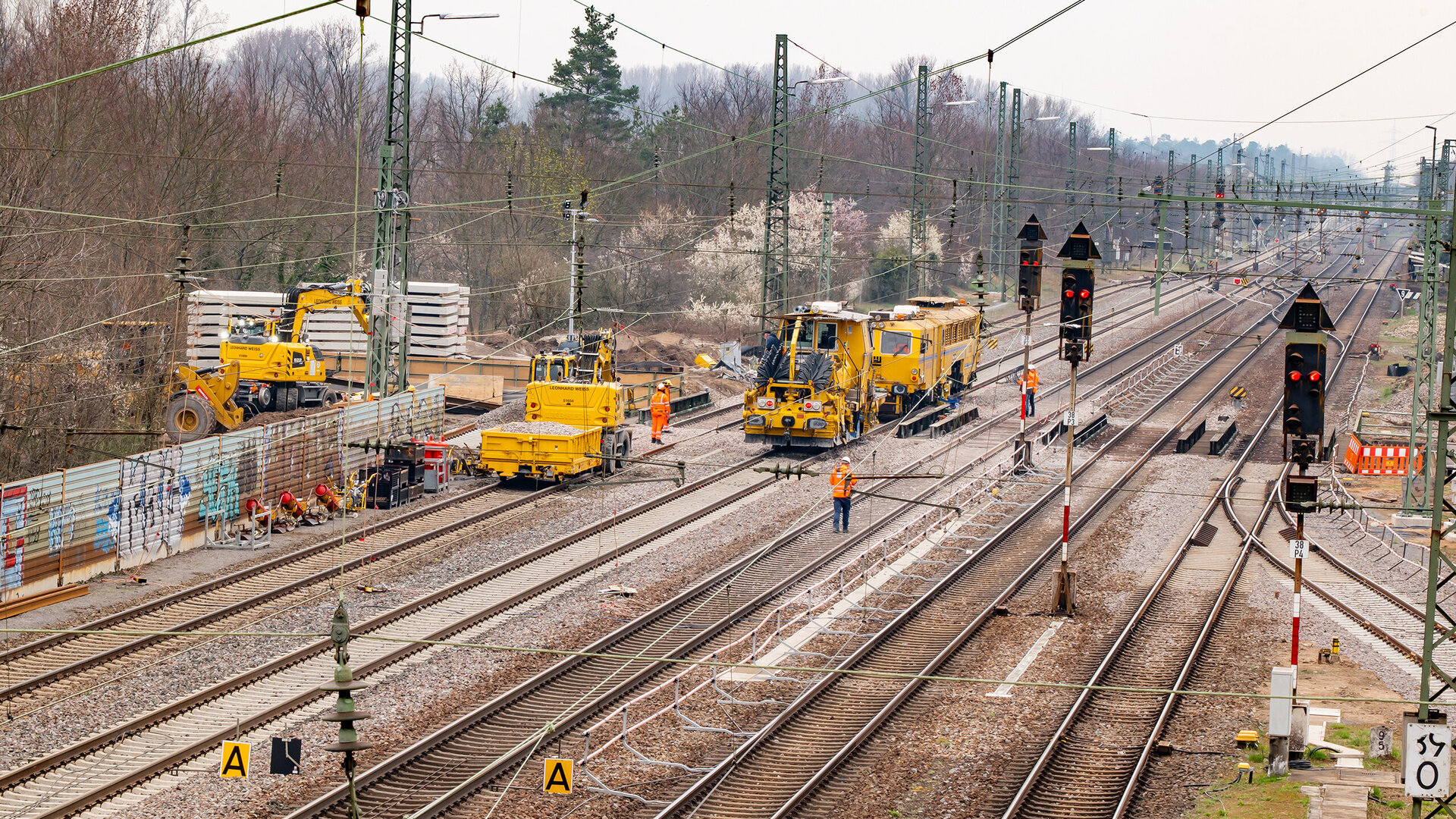 Several yellow machines are working on the tracks at a construction site, while workers in orange clothing are busy at work.