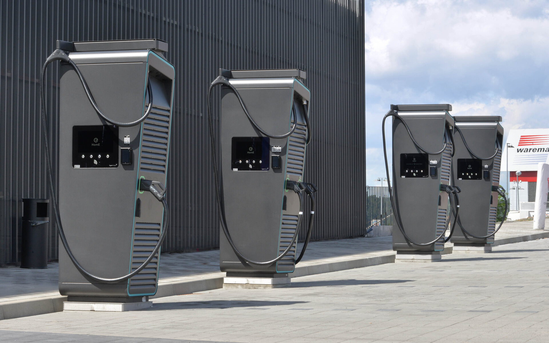 Vier moderne Ladestationen für Elektroautos stehen auf einem gepflasterten Platz, umgeben von einer hohen Wand.