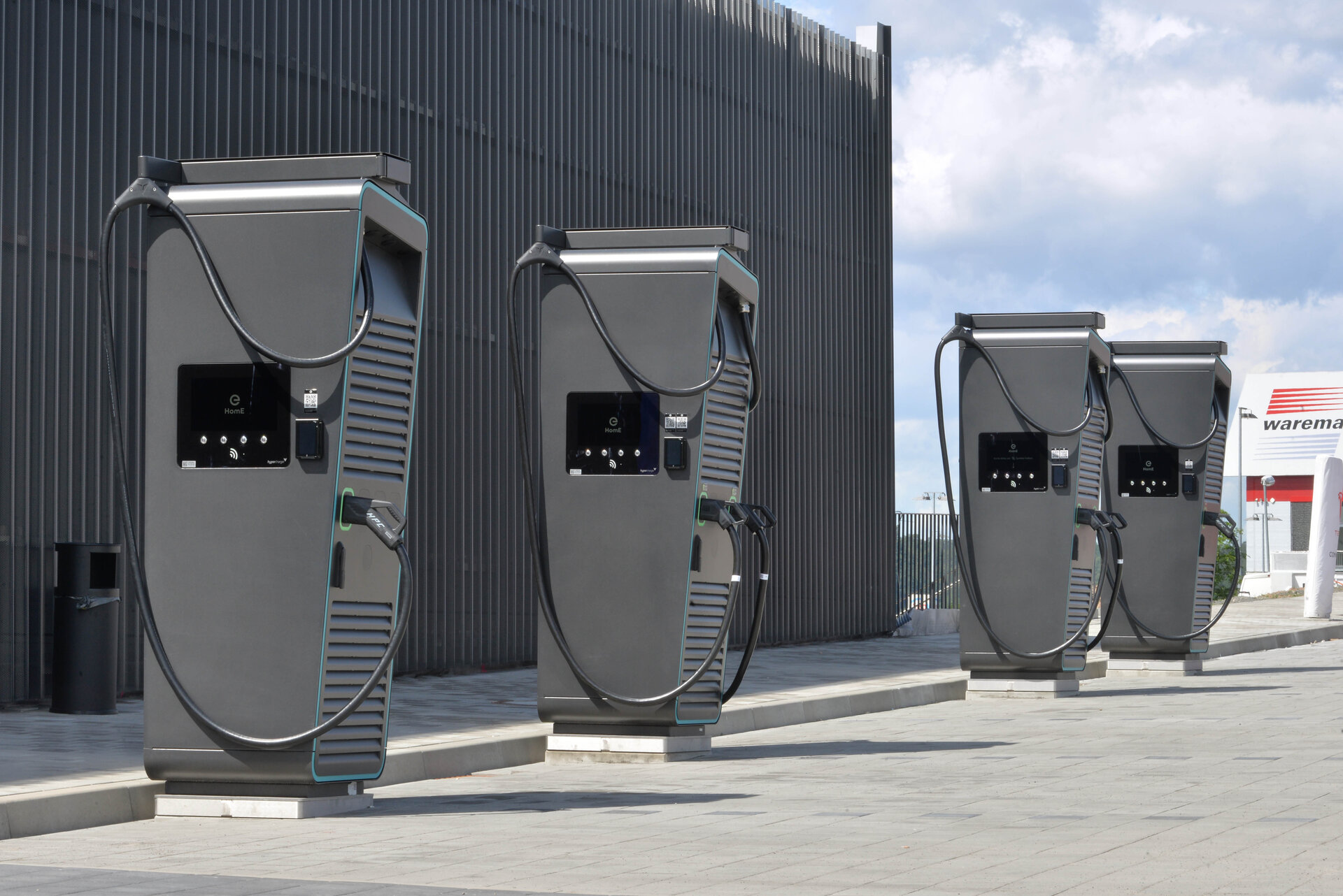 Vier moderne Ladestationen für Elektroautos stehen auf einem gepflasterten Platz, umgeben von einer hohen Wand.