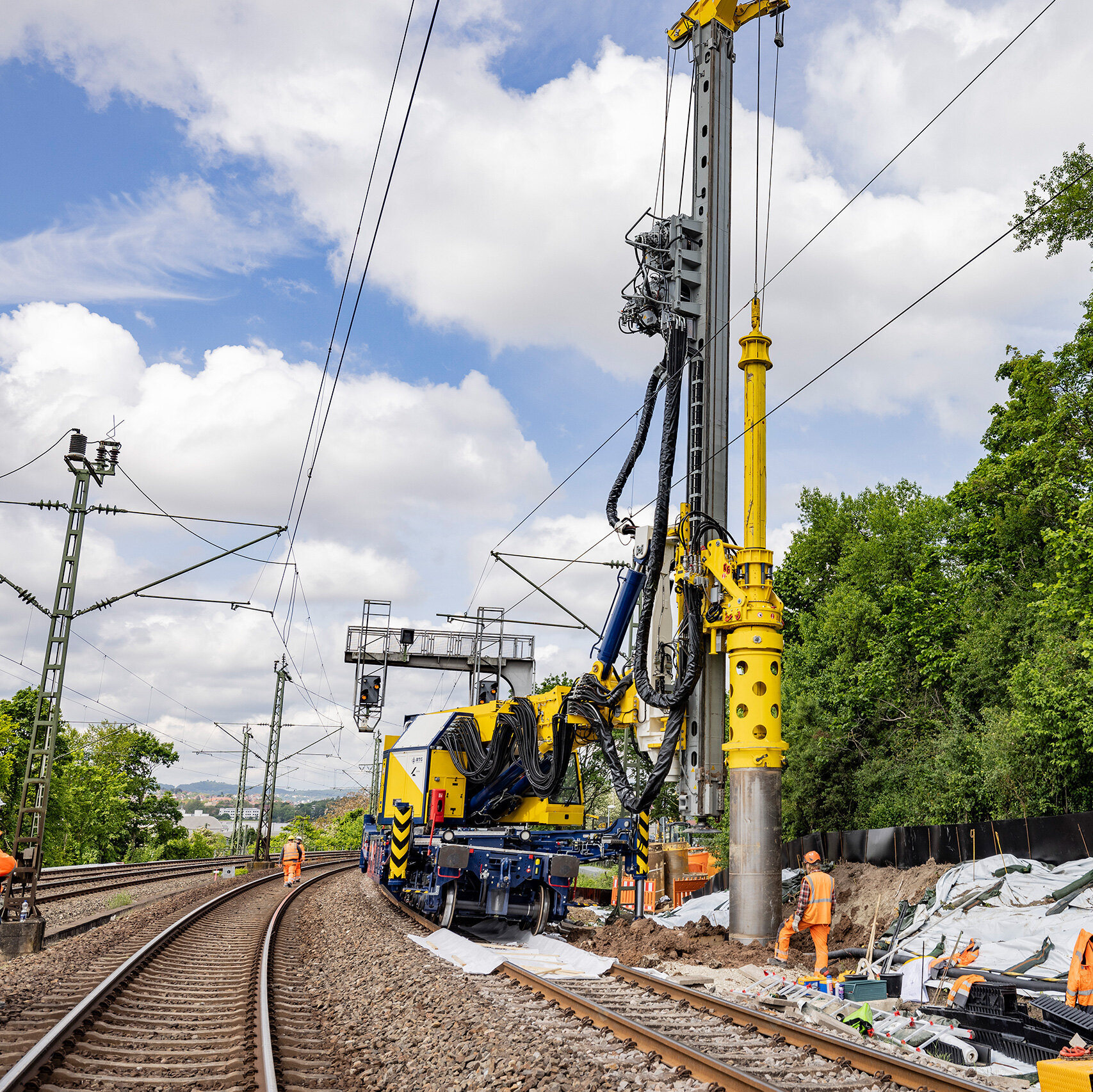 Die Szene zeigt eine Ramme, die an den Gleisen arbeitet, umgeben von Bäumen und einem klaren Himmel mit Wolken.