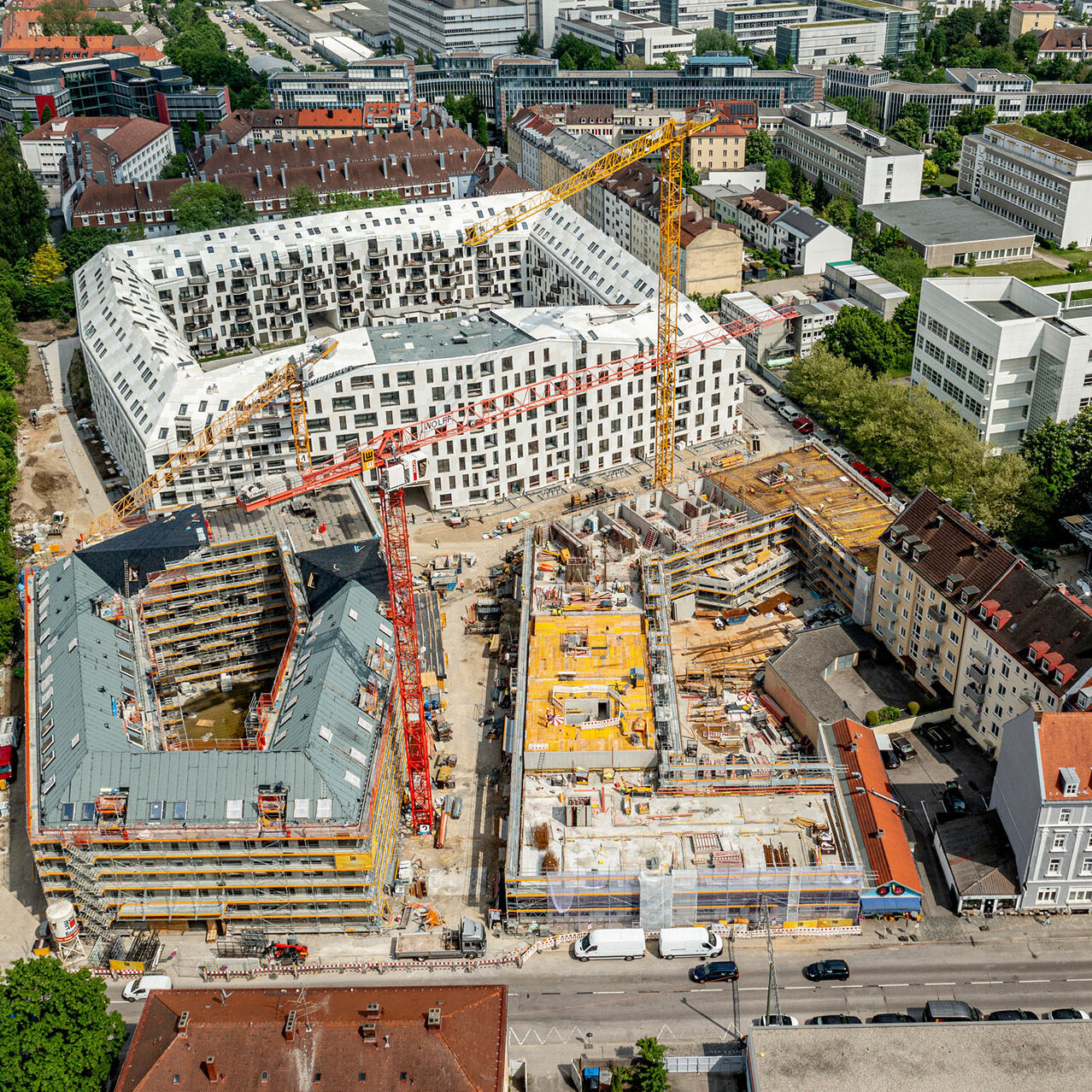 Luftansicht einer aktiven Baustelle mit verschiedenen Bauphasen, umgeben von urbaner Landschaft und grünen Flächen.