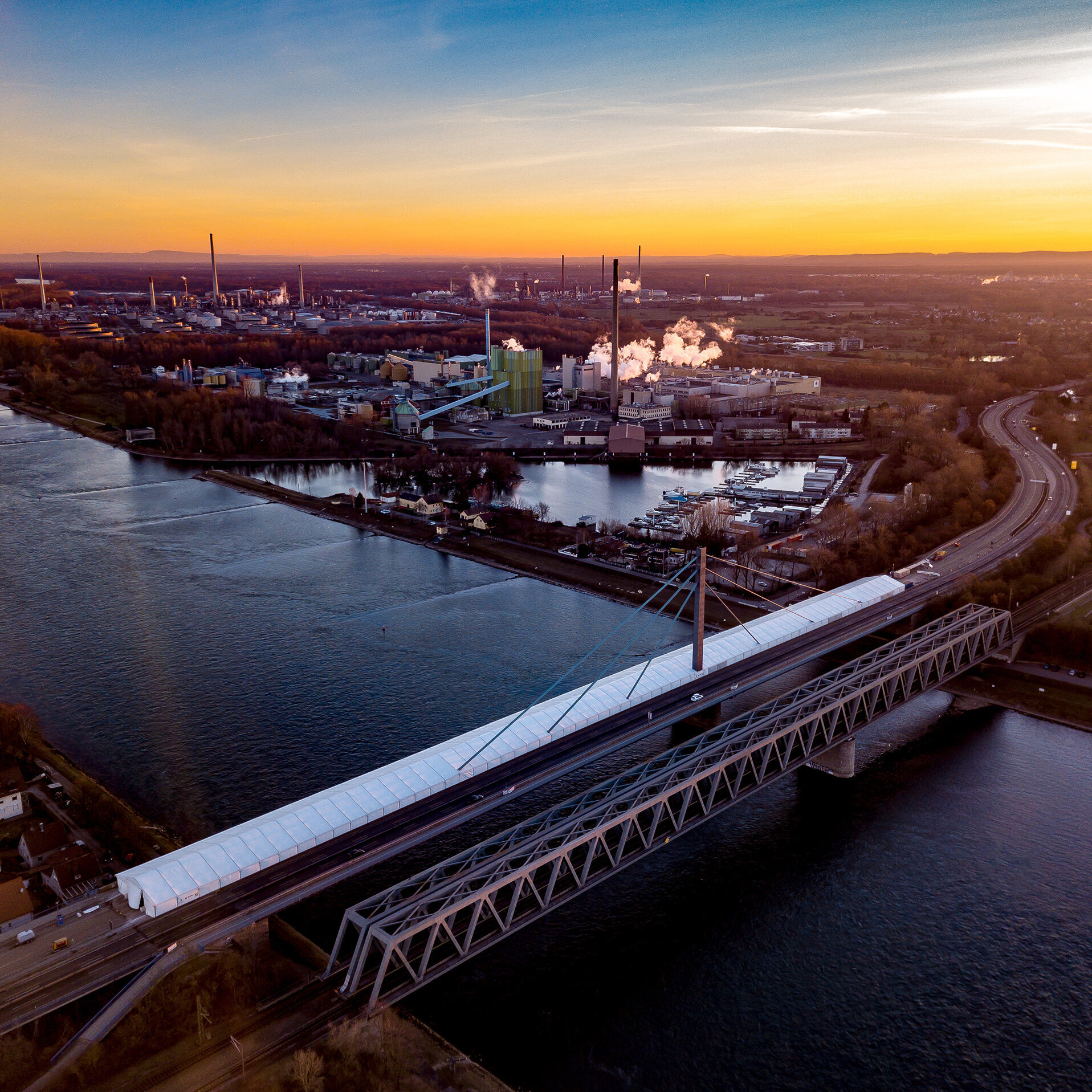 Sonnenuntergang über einer industriellen Umgebung, wo eine moderne Brücke den Verkehr über den Fluss leitet.