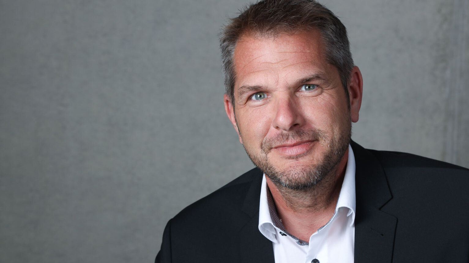 Vor einem schlichten Hintergrund präsentiert sich der Mann mit einem freundlichen Lächeln und einem modischen, formellen Outfit.