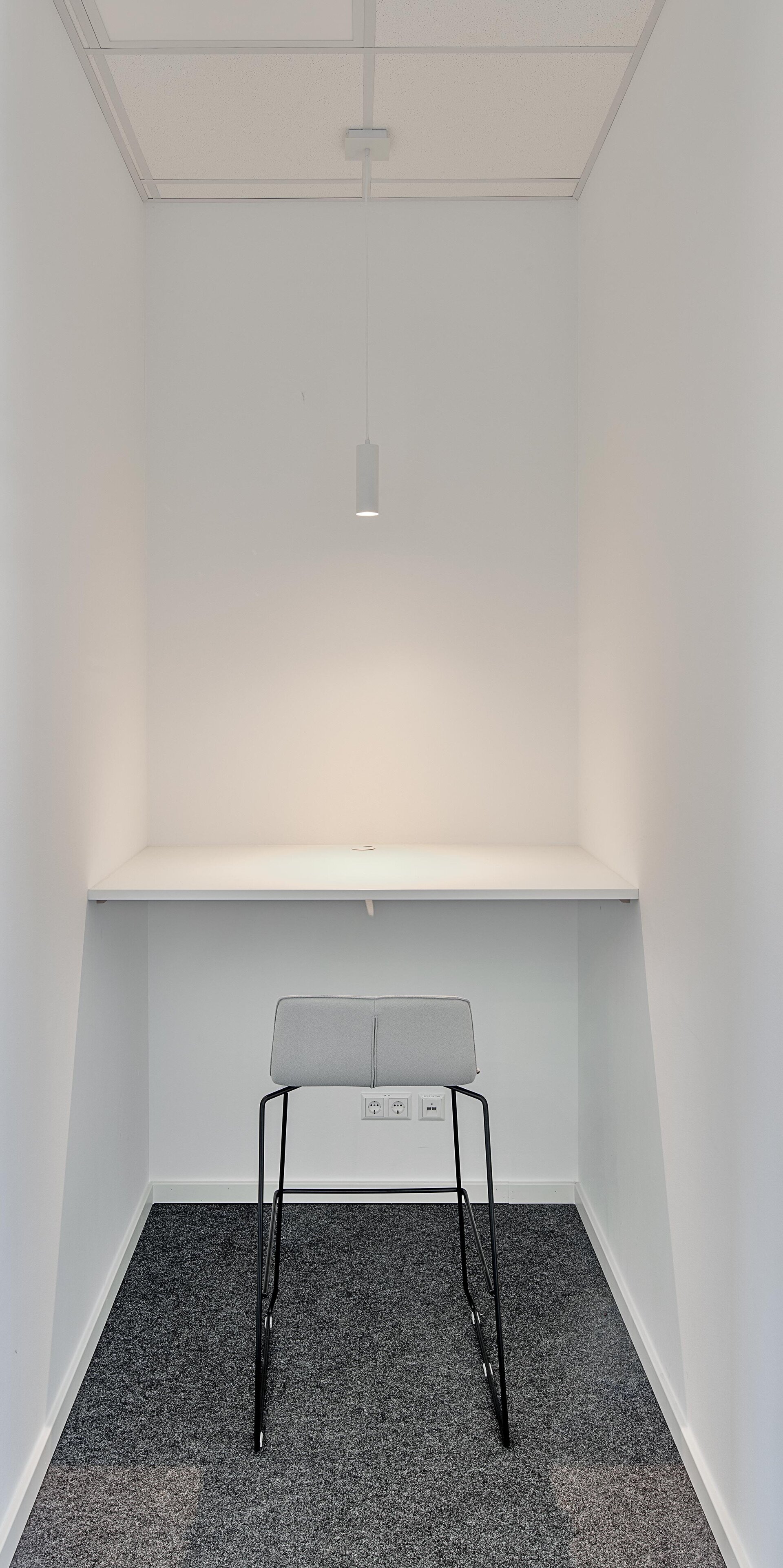 Ein minimalistischer Raum mit einem grauen Hocker und einem schlichten weißen Tisch, beleuchtet von einer modernen Lampe.