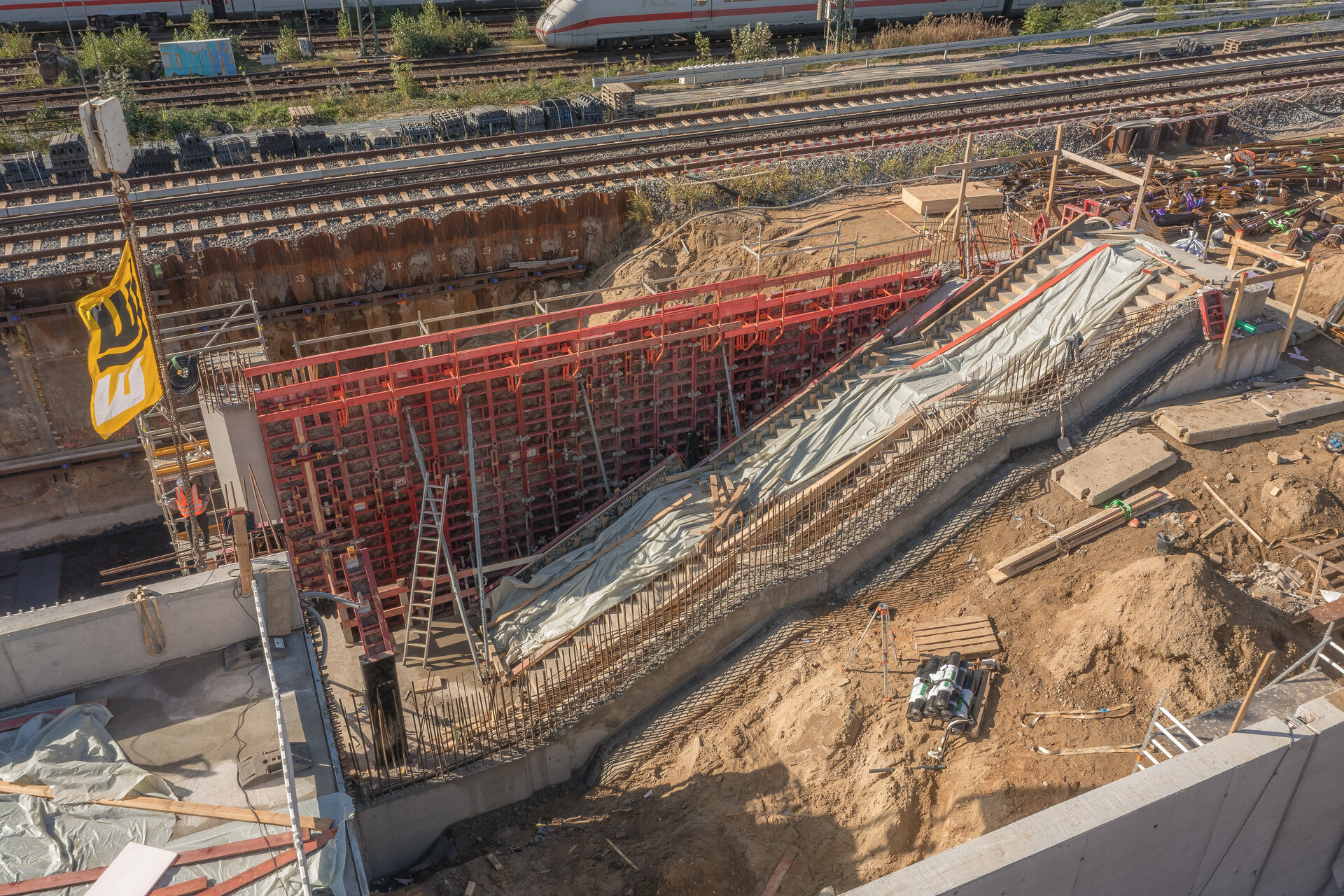Eine Baustelle mit einem großen, roten Schalungsgerüst, das für den Bau einer Rampe vorbereitet ist, umgeben von Erde und Werkzeugen.