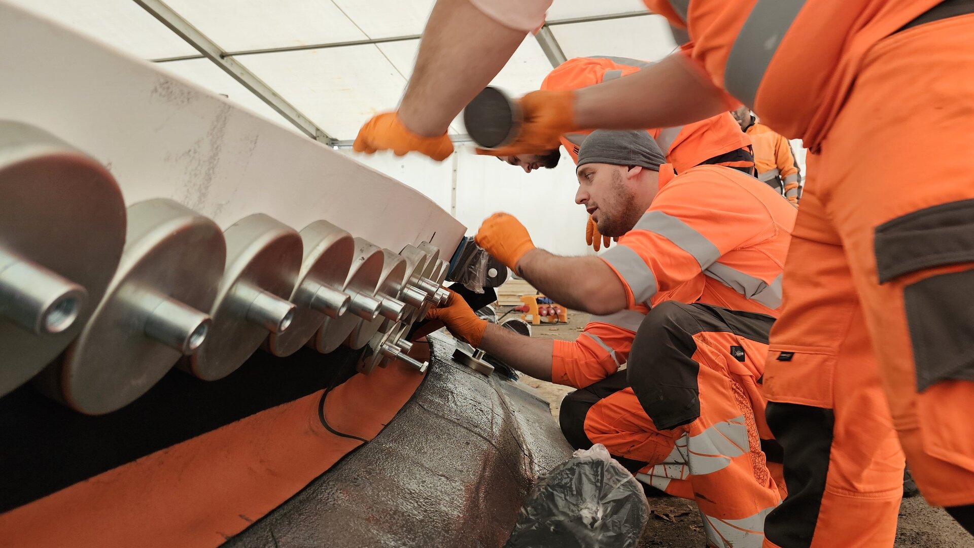 Mehrere Arbeiter in orangefarbener Arbeitskleidung arbeiten an der Dehnfugenabdichtung an einem großen Gerät unter einem Zelt.