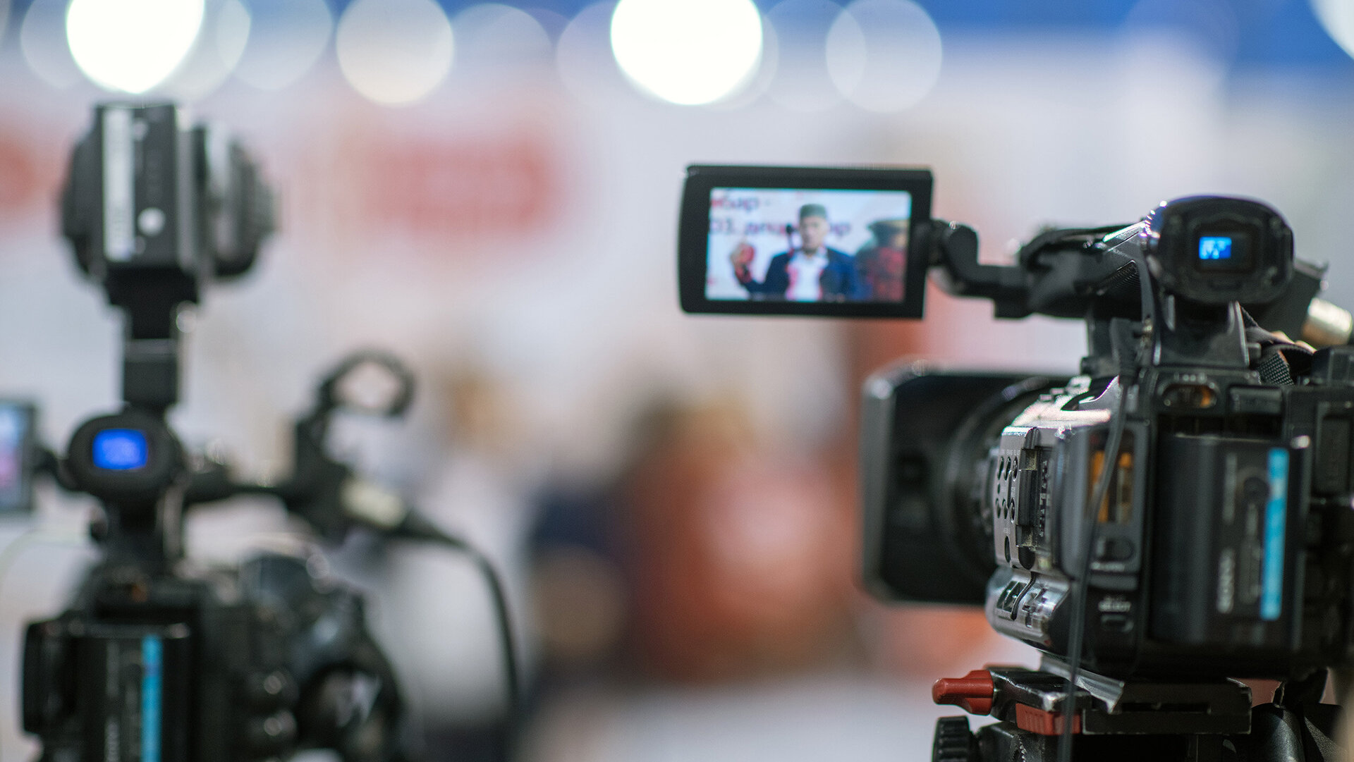 Im Bild sind zwei Videokameras zu sehen, die auf einen Redner fokussiert sind, der in einem hellen Raum spricht.