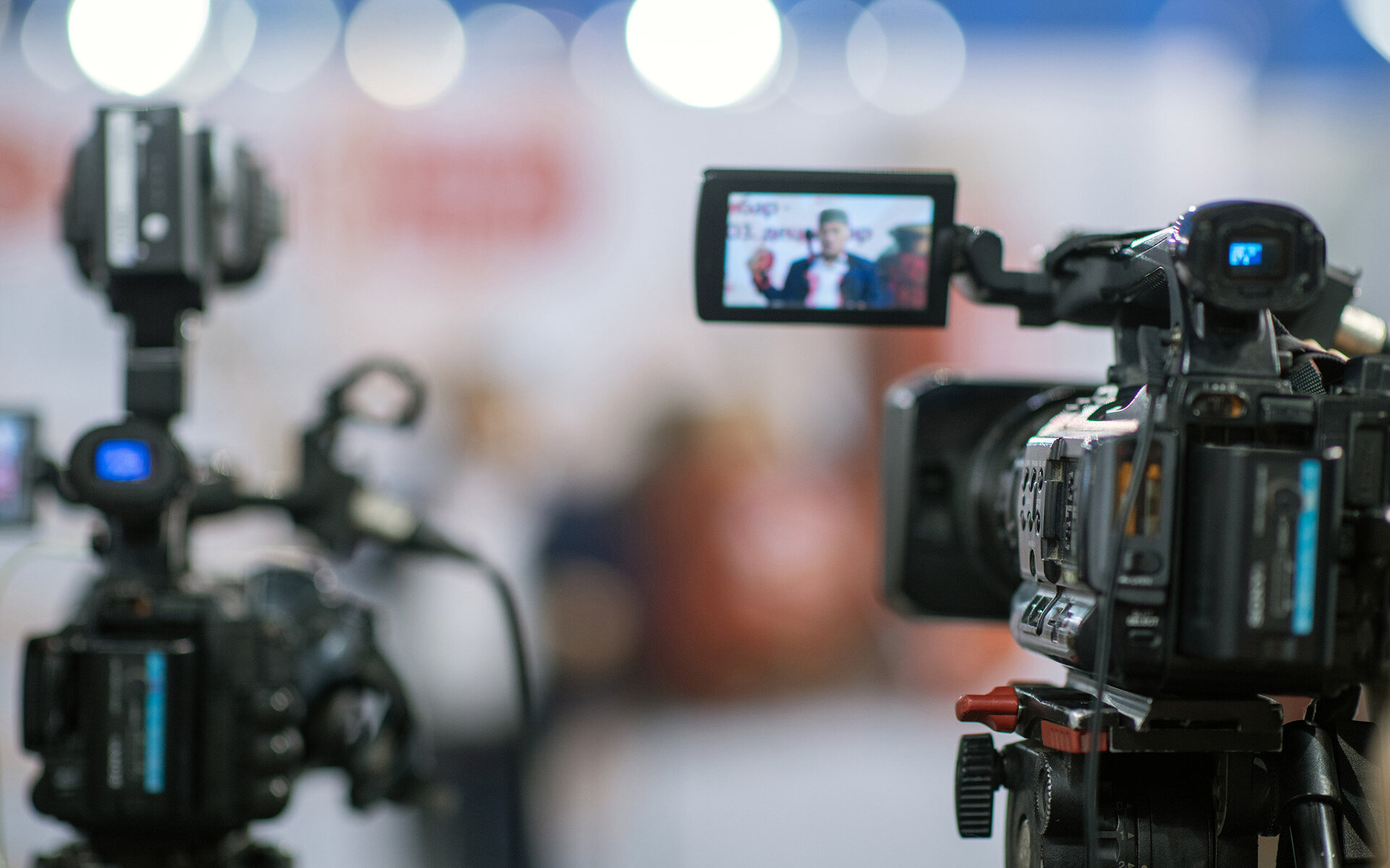 Im Bild sind zwei Videokameras zu sehen, die auf einen Redner fokussiert sind, der in einem hellen Raum spricht.