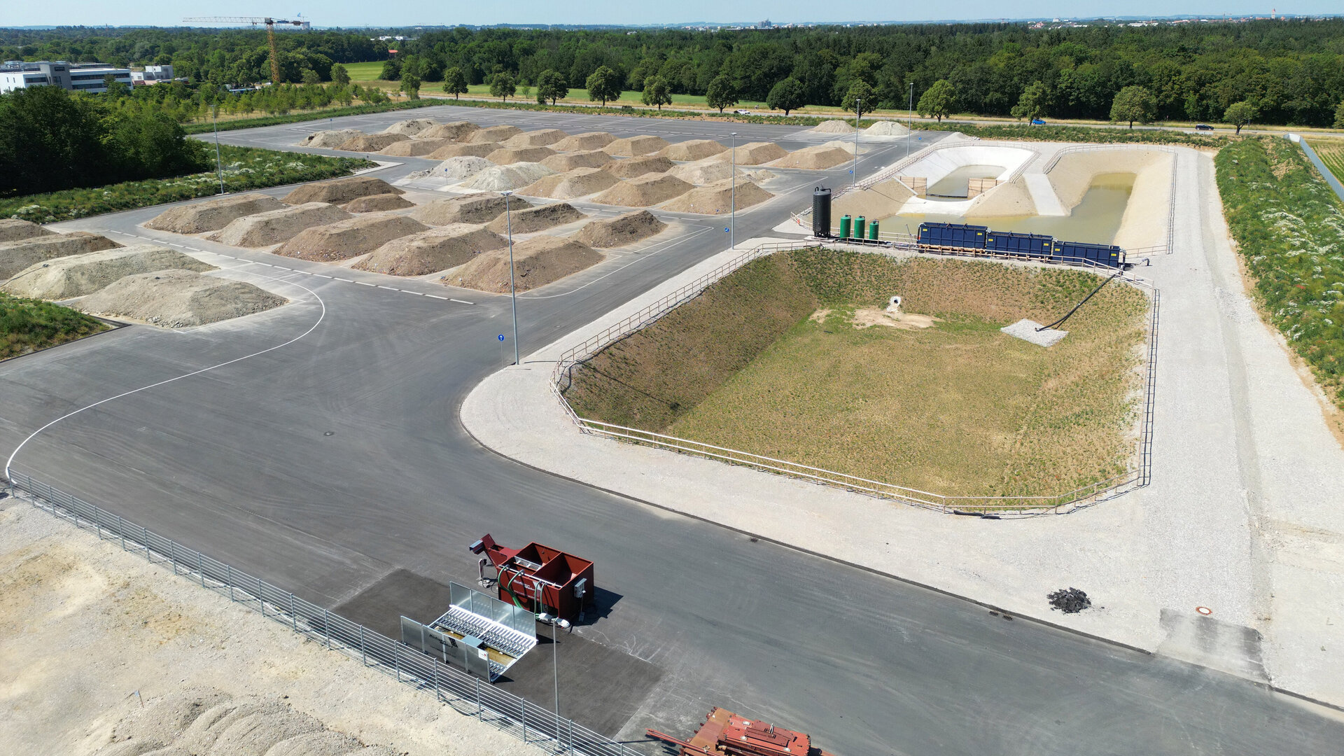 Die Szene zeigt eine große Fläche mit verschiedenen Erdhaufen, umgeben von einer asphaltierten Straße und grünen Bäumen.