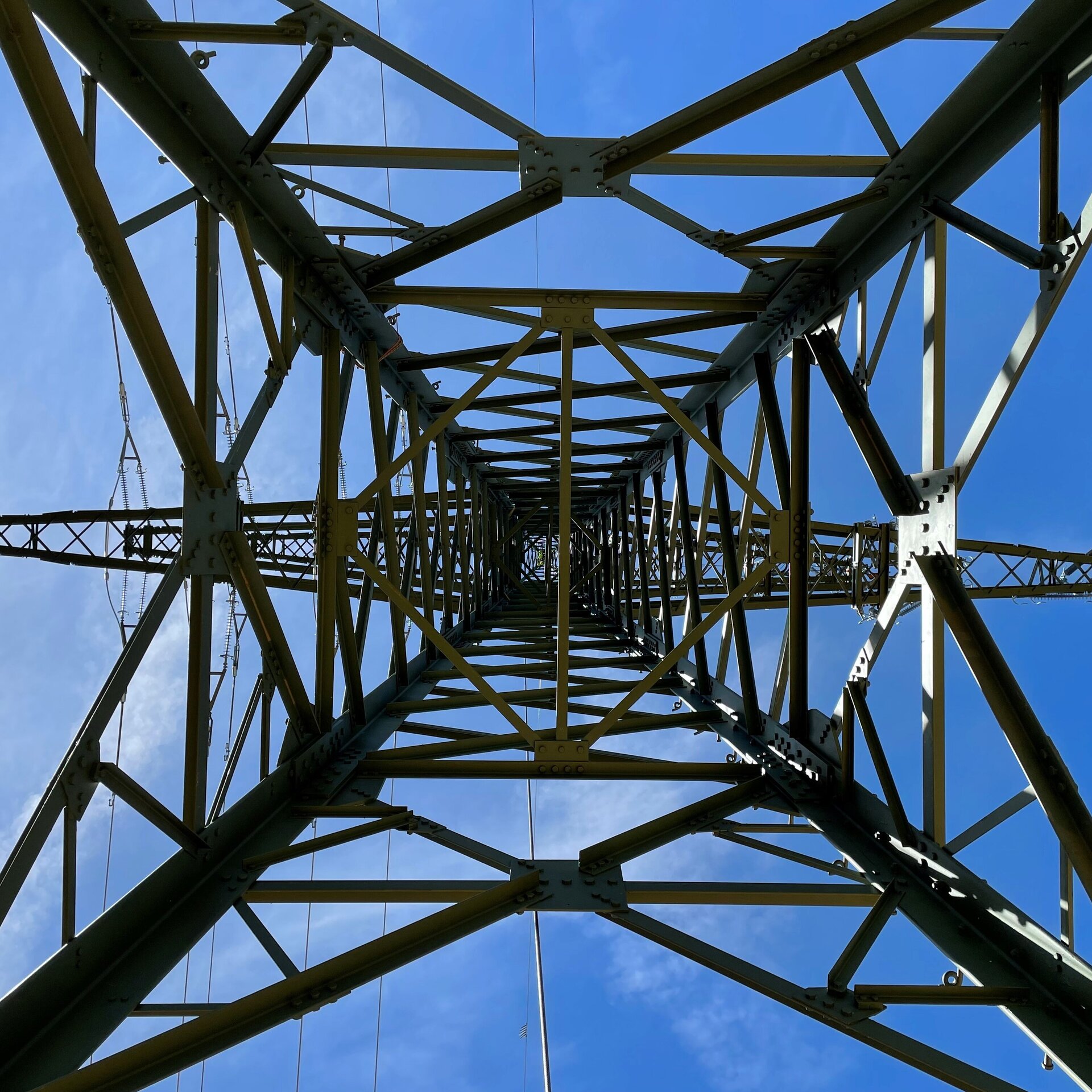 Die komplexe Konstruktion eines Strommasts wird von unten betrachtet, umgeben von einem hellen, wolkenlosen Himmel.