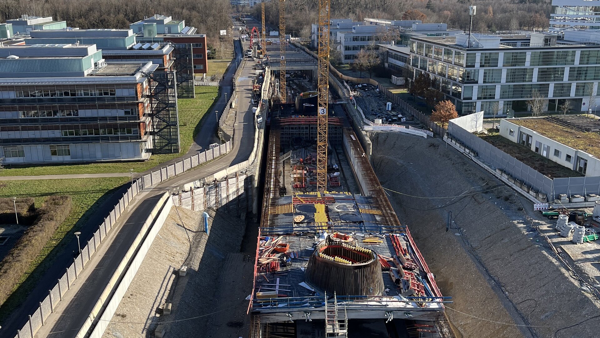 Eine Baustelle mit einem großen Kran, der über einen tiefen Graben schwebt, umgeben von modernen Gebäuden und einem blauen Himmel.