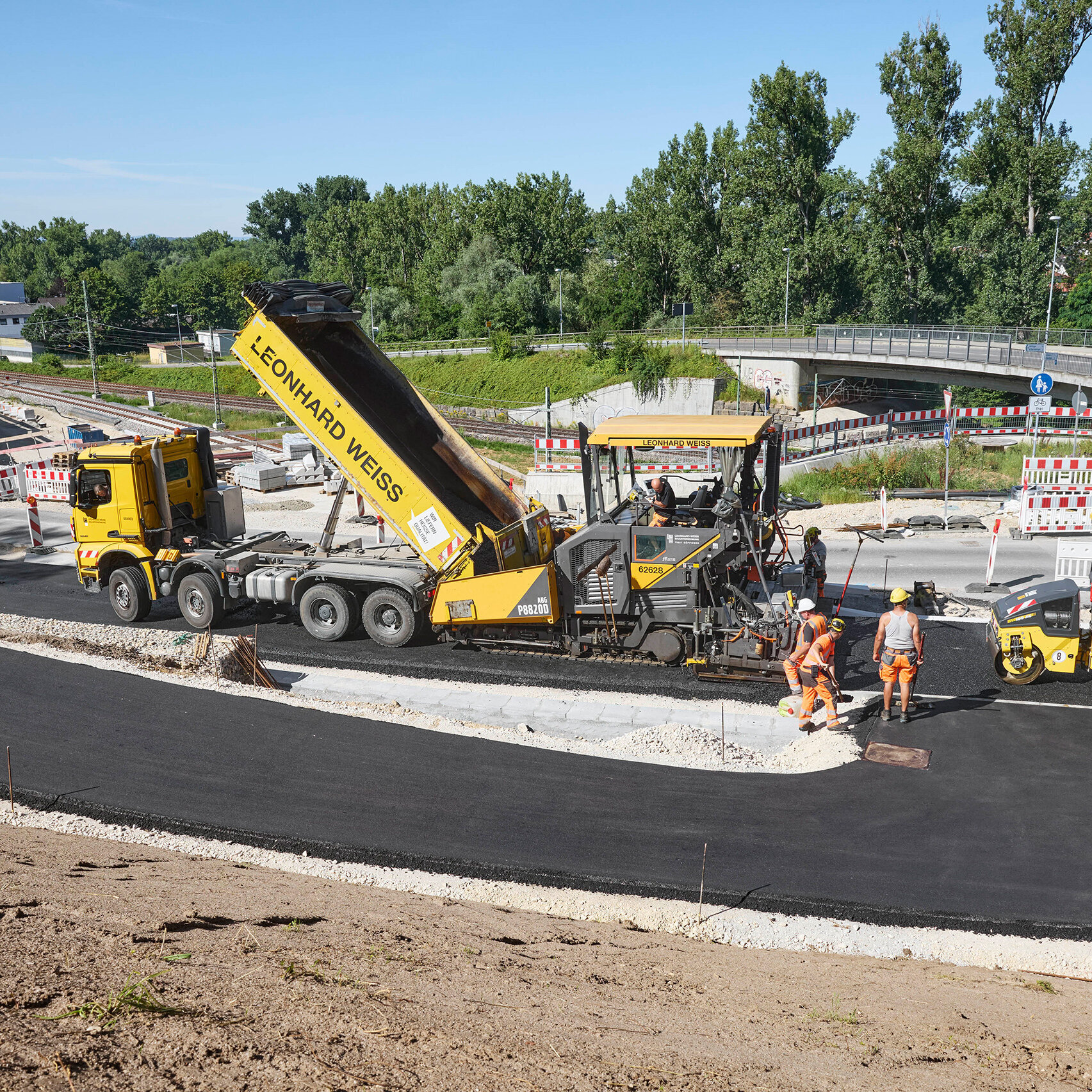 Eine Baustelle mit mehreren Arbeitern, die an der Asphaltierung einer Straße mit schweren Maschinen arbeiten.