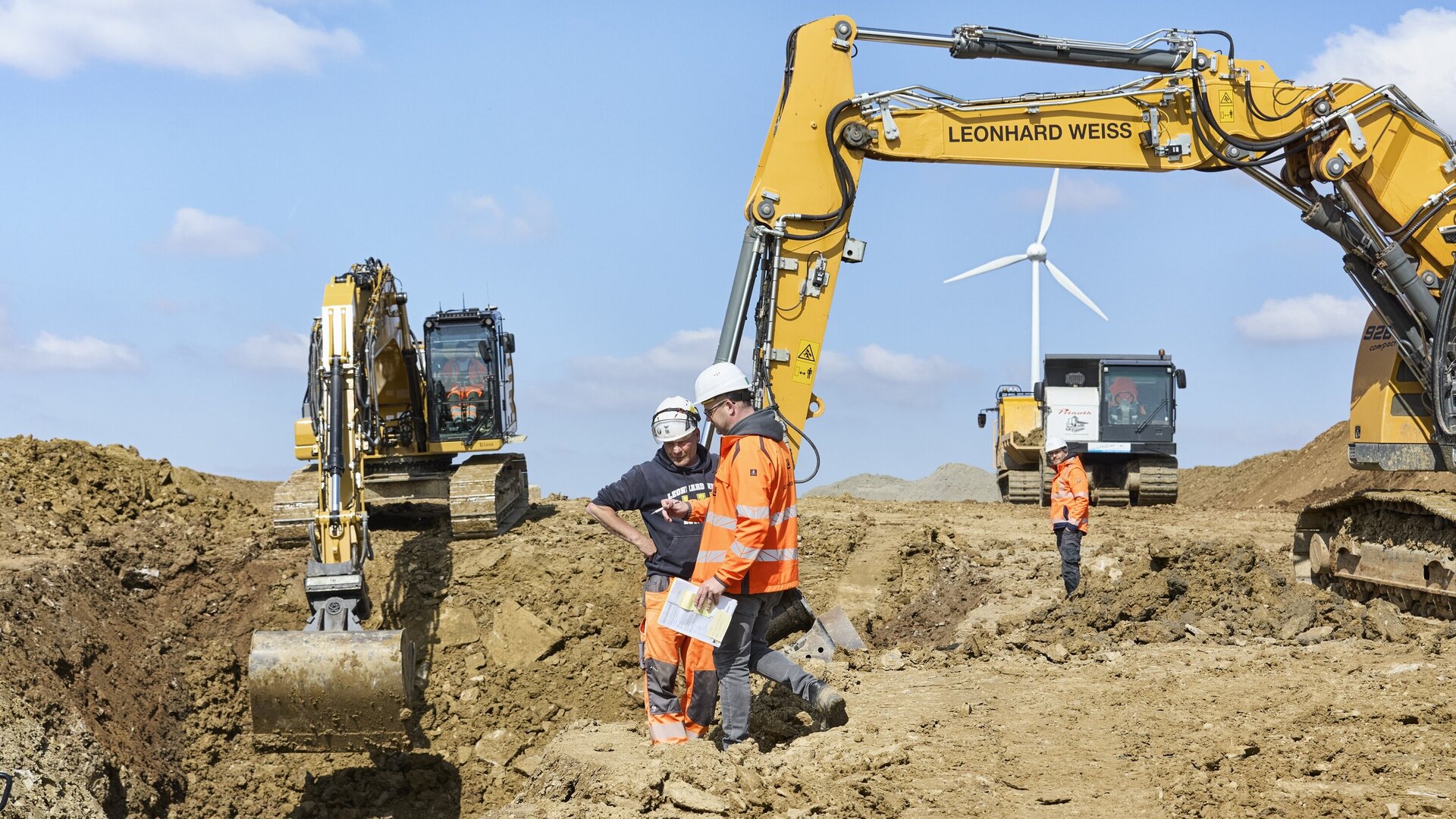 Zwei Männer in Sicherheitskleidung stehen auf unebenem Boden und besprechen die Arbeiten, während Bagger im Hintergrund graben.