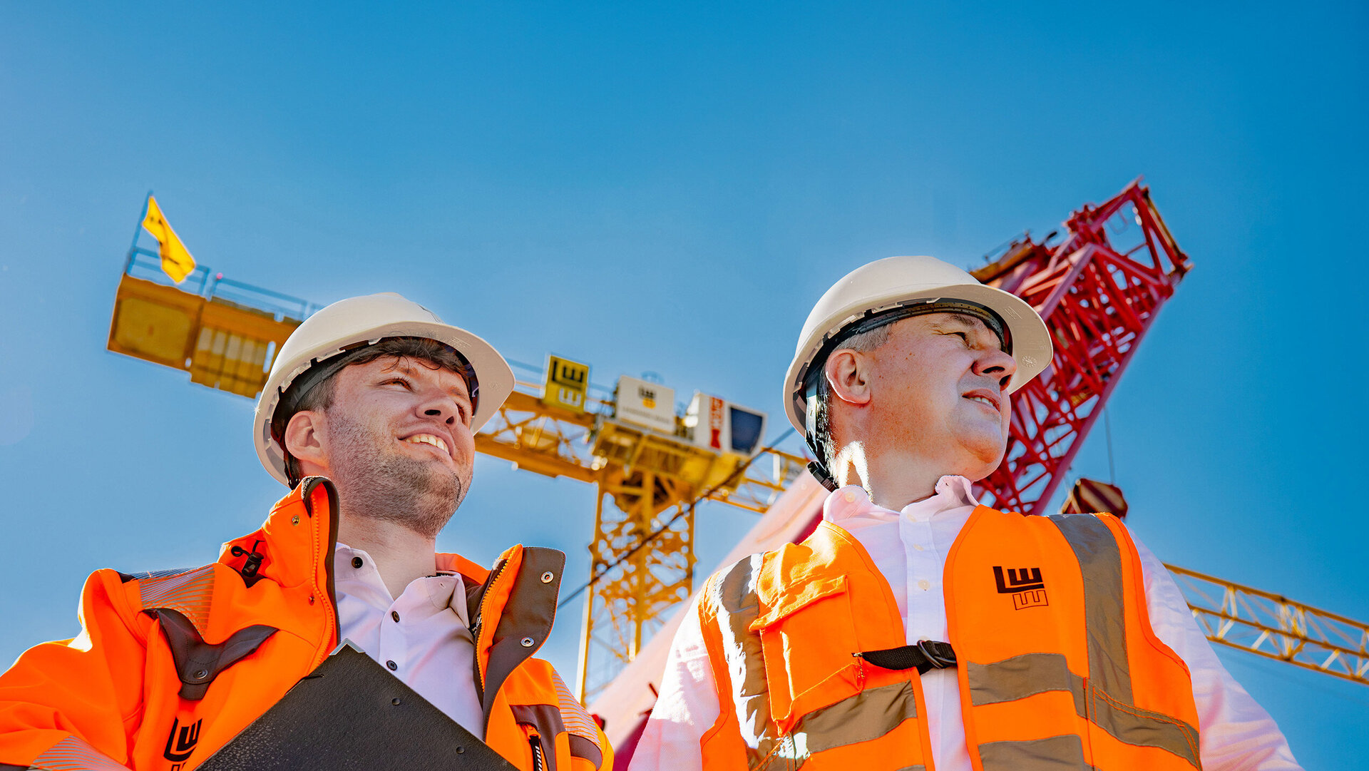 Zwei Männer in leuchtenden Sicherheitswesten, einer mit einer Arbeitsmappe in der Hand, stehen unter einem blauen Himmel, während im Hintergrund ein Kran zu sehen ist.