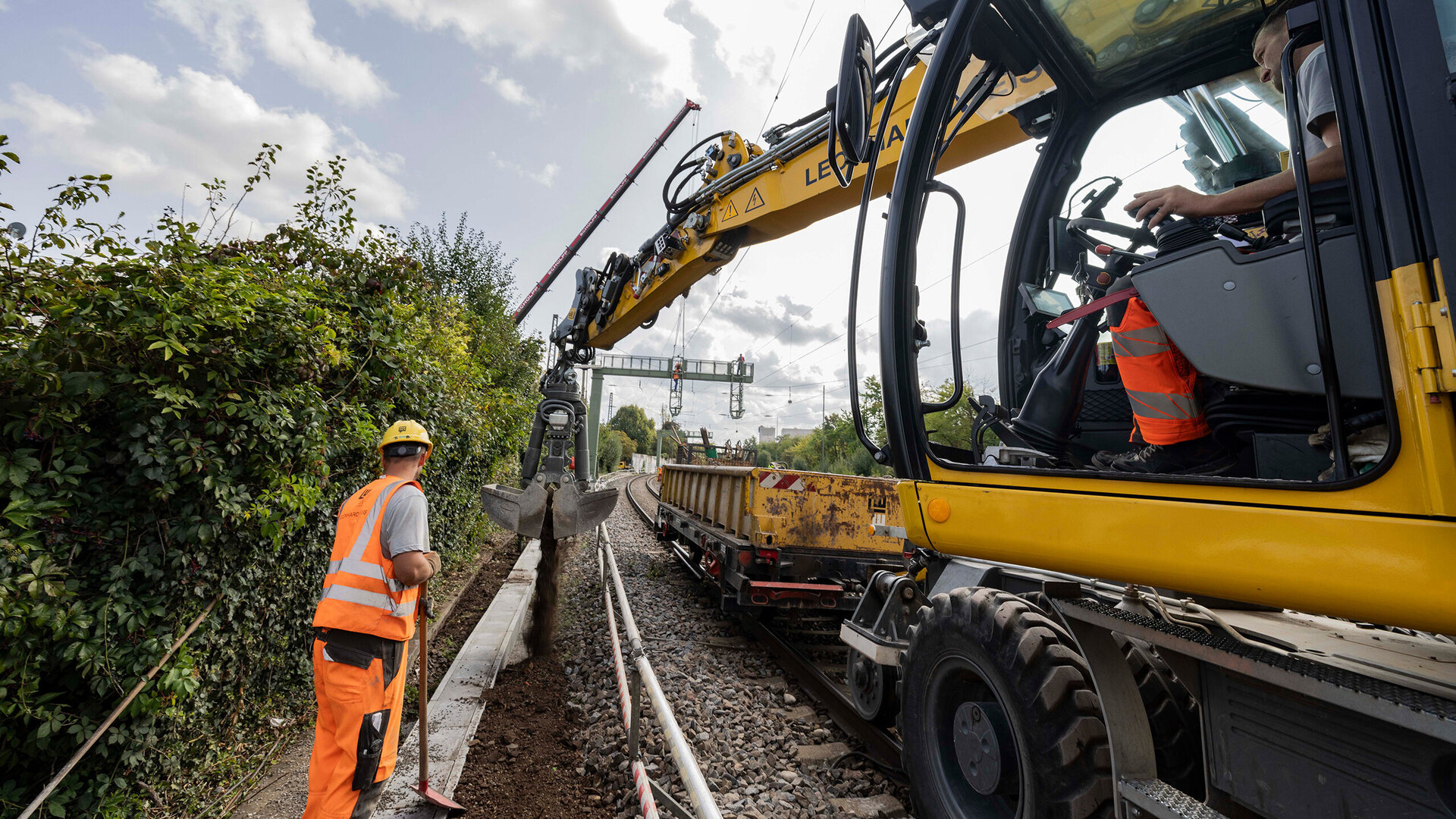 Ein gelber Schienenbagger arbeitet am Gleis-Kabelkanal, während ein Bauarbeiter in orangefarbener Kleidung die Arbeiten überwacht.