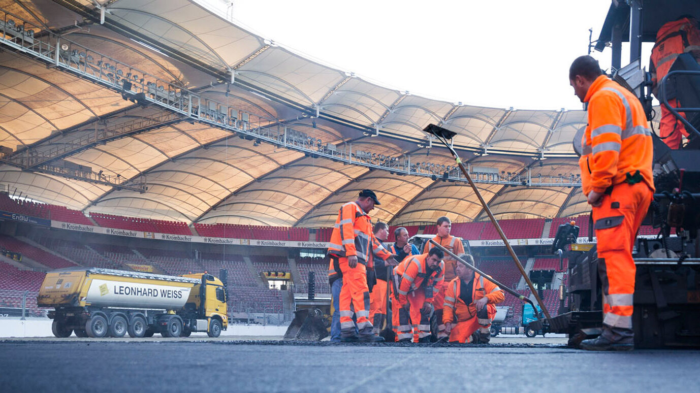 In einem Stadion arbeiten mehrere Bauarbeiter in orangefarbener Kleidung an der Asphaltierung des Spielfelds.