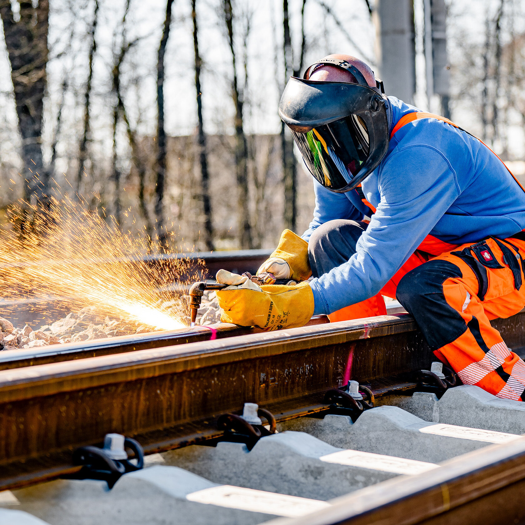 Ein Mann in orangefarbener Arbeitskleidung schweißt an den Gleisen, während Funken sprühen und die Bäume im Hintergrund sichtbar sind.