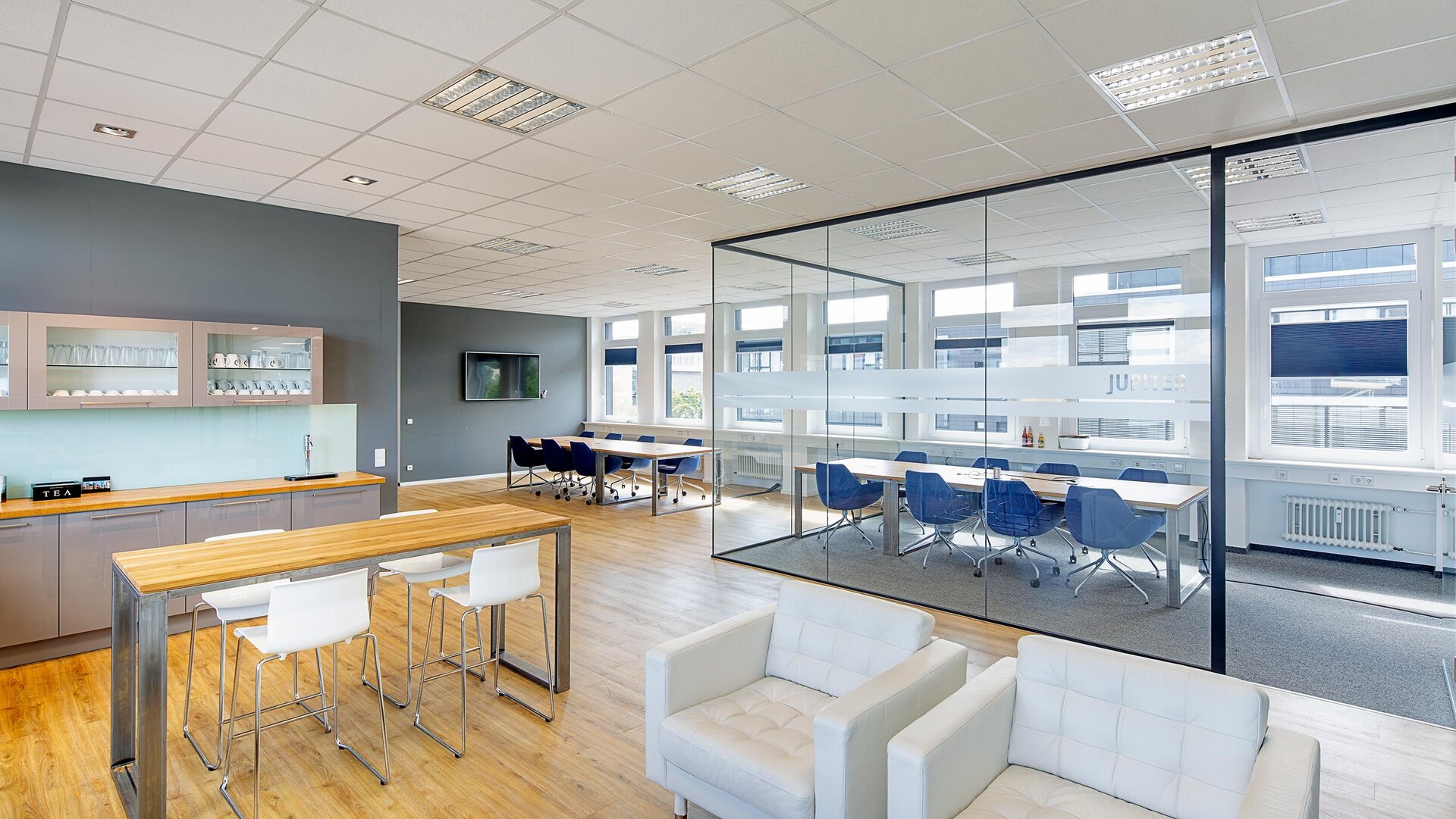 Ein modernes Büro mit einer hellen Küche, einem langen Tisch und blauen Stühlen, ideal für Besprechungen und Pausen.