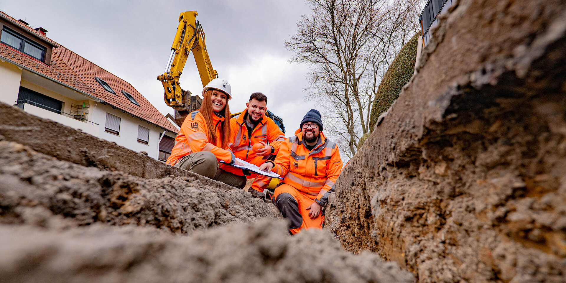 Drei Bauarbeiter, zwei Männer und eine Frau mit einem Plan vor sich, in leuchtend orangefarbener Arbeitskleidung lächeln fröhlich in einem Graben, während ein Bagger im Hintergrund steht.