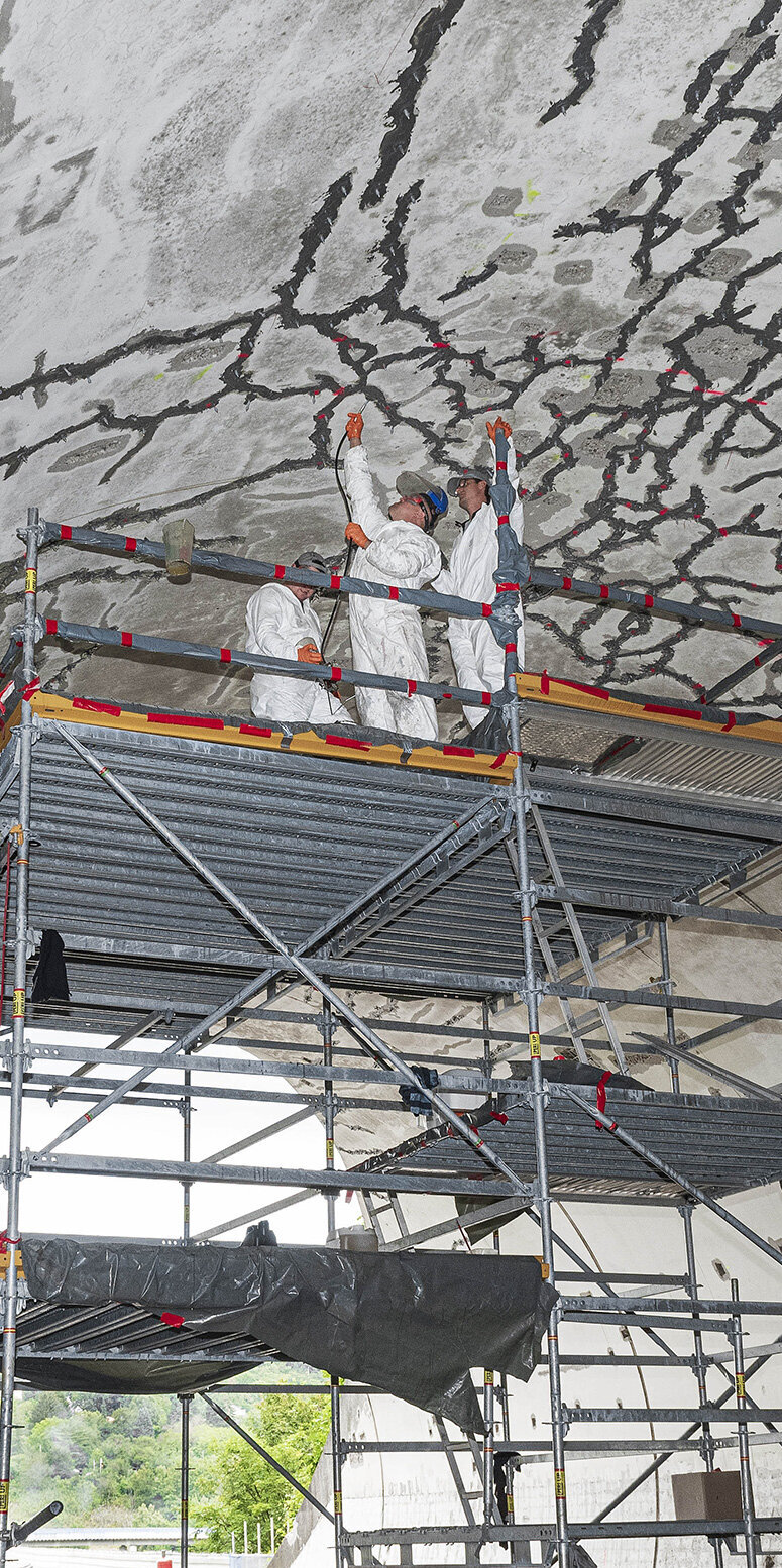 Arbeiter in Schutzanzügen inspizieren die Risse an der Decke eines Tunnels, während sie auf einem Gerüst stehen.