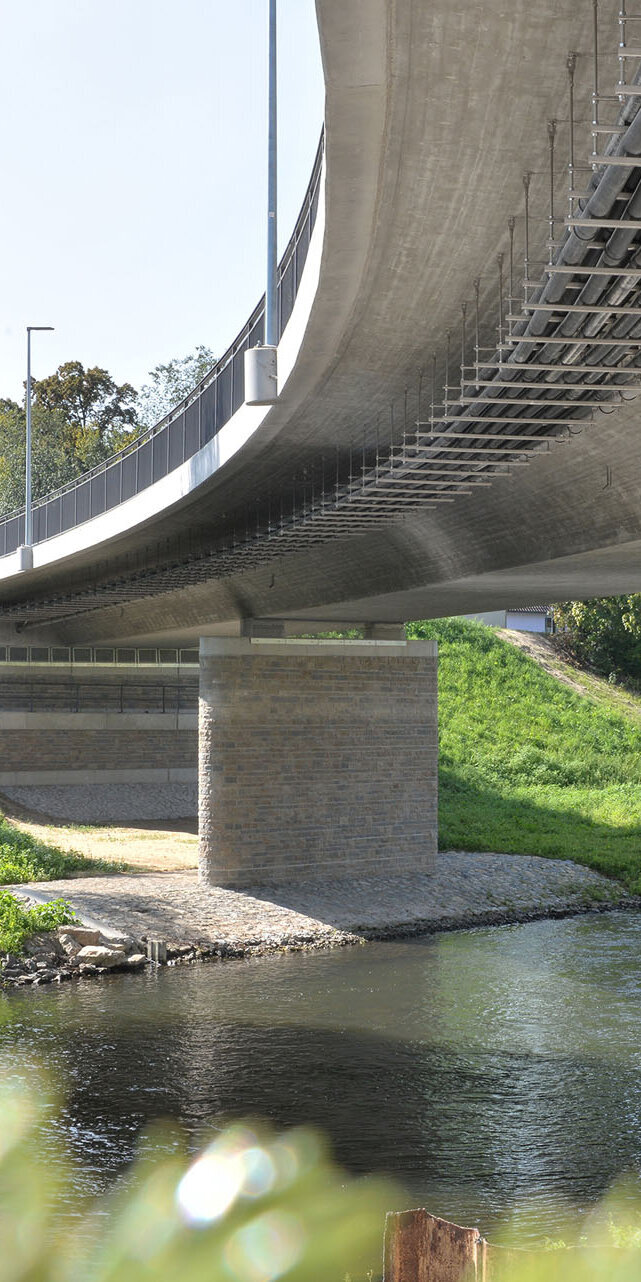 Ein Blick auf die Unterseite einer Brücke, die über einen Fluss führt, mit massiven Pfeilern und einer grünen Uferlandschaft.