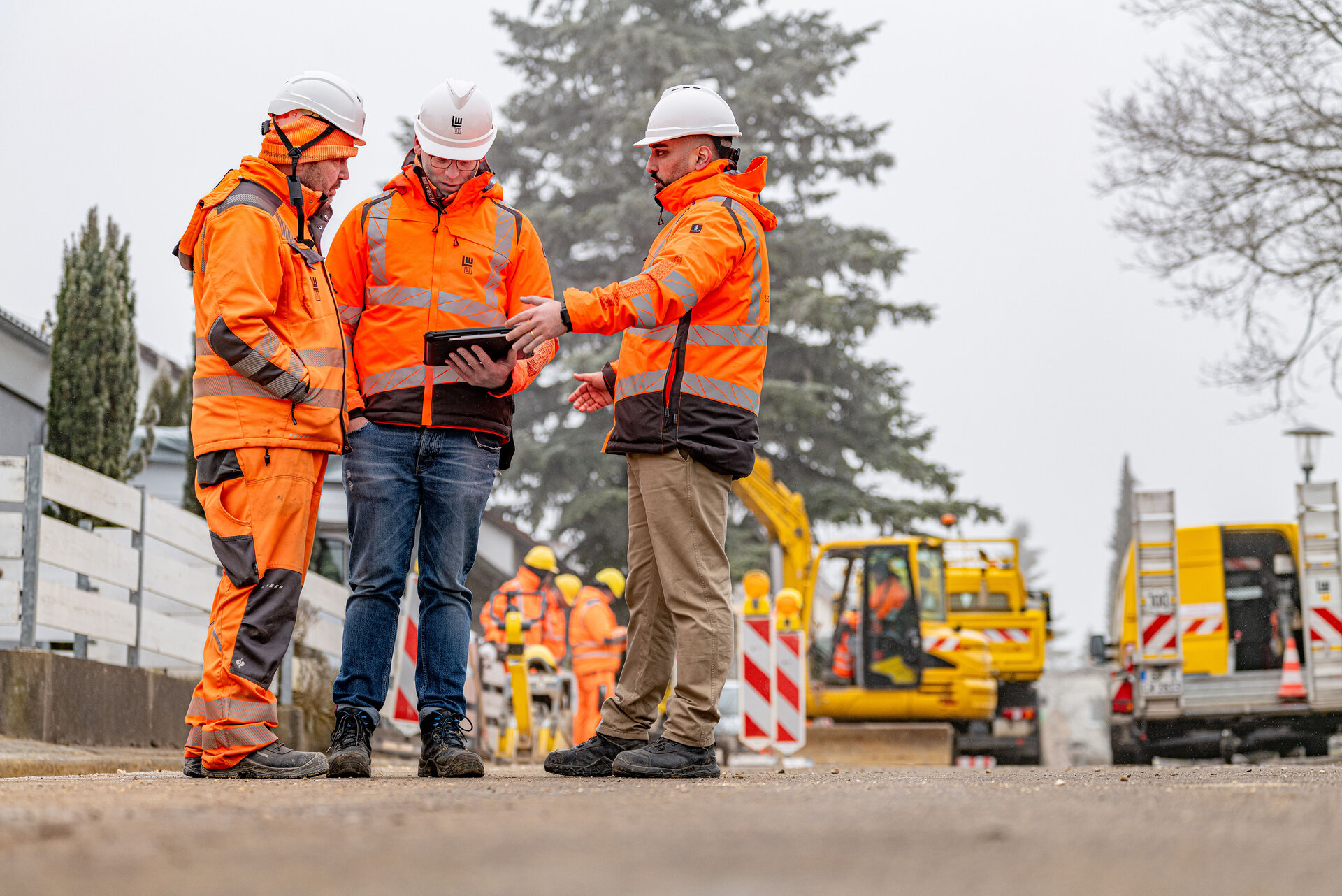 Drei Bauarbeiter in leuchtend orangefarbener Kleidung besprechen Pläne auf einem Tablet, während im Hintergrund Maschinen und weitere Arbeiter arbeiten.