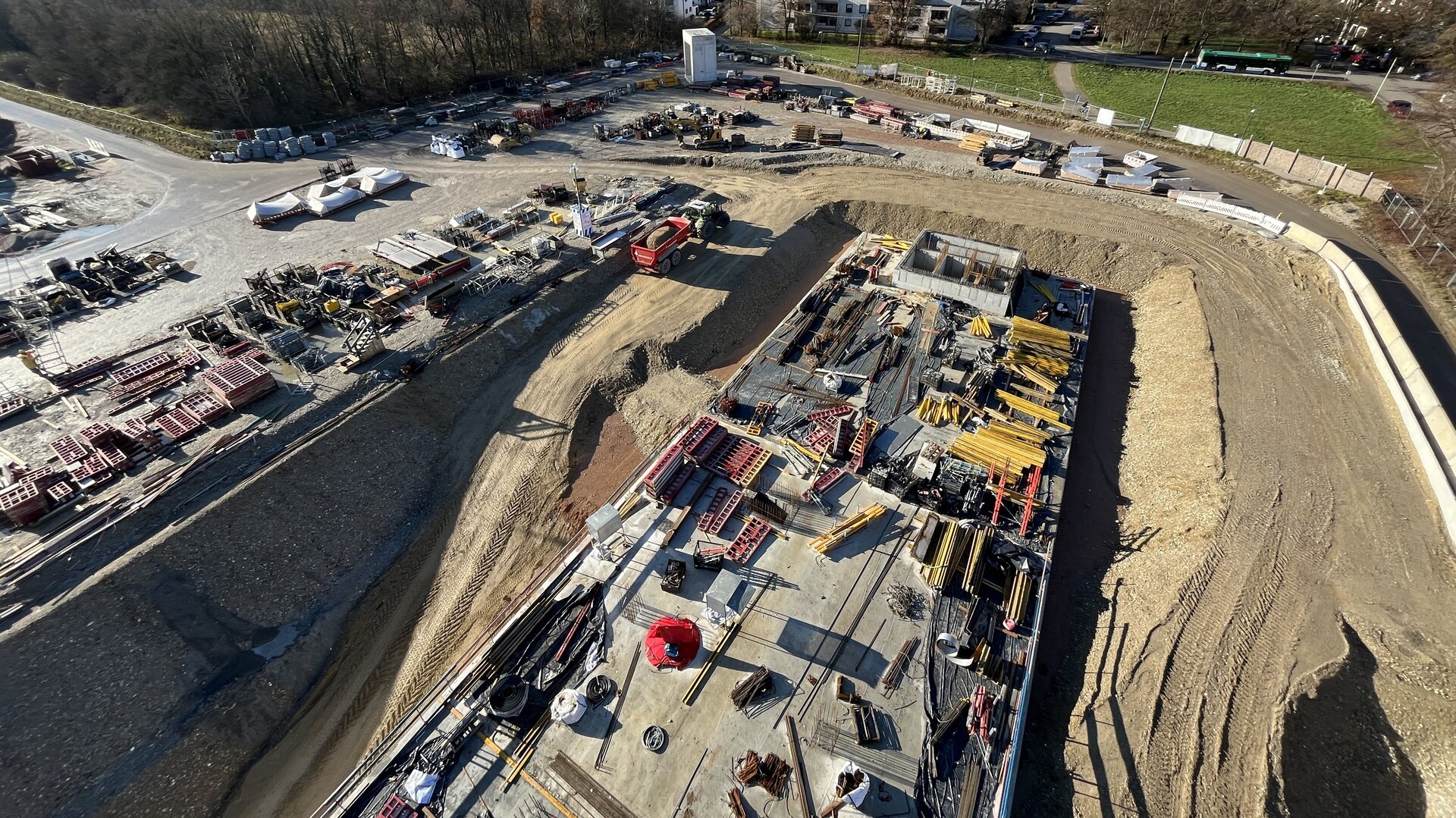 Luftaufnahme einer Baustelle mit verschiedenen Maschinen und Materialien, im Hintergrund eine Stadtlandschaft mit Bäumen und einem klaren blauen Himmel.