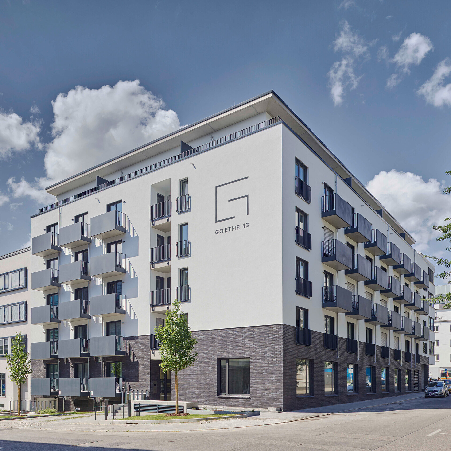 Ein modernes Gebäude mit mehreren Balkonen und großen Fenstern, umgeben von Bäumen und einem klaren blauen Himmel.