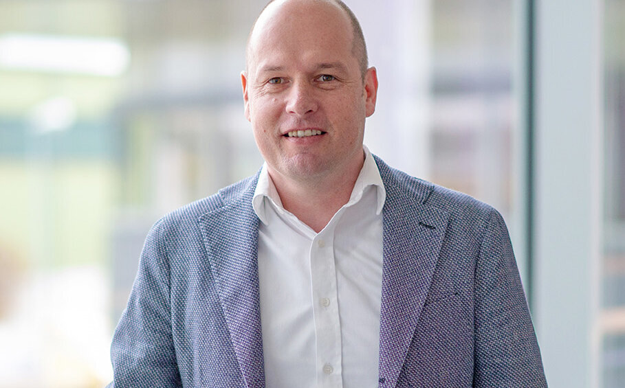 Mit einem warmen Lächeln präsentiert sich ein Mann in einem stilvollen Büro, das von natürlichem Licht durchflutet wird.